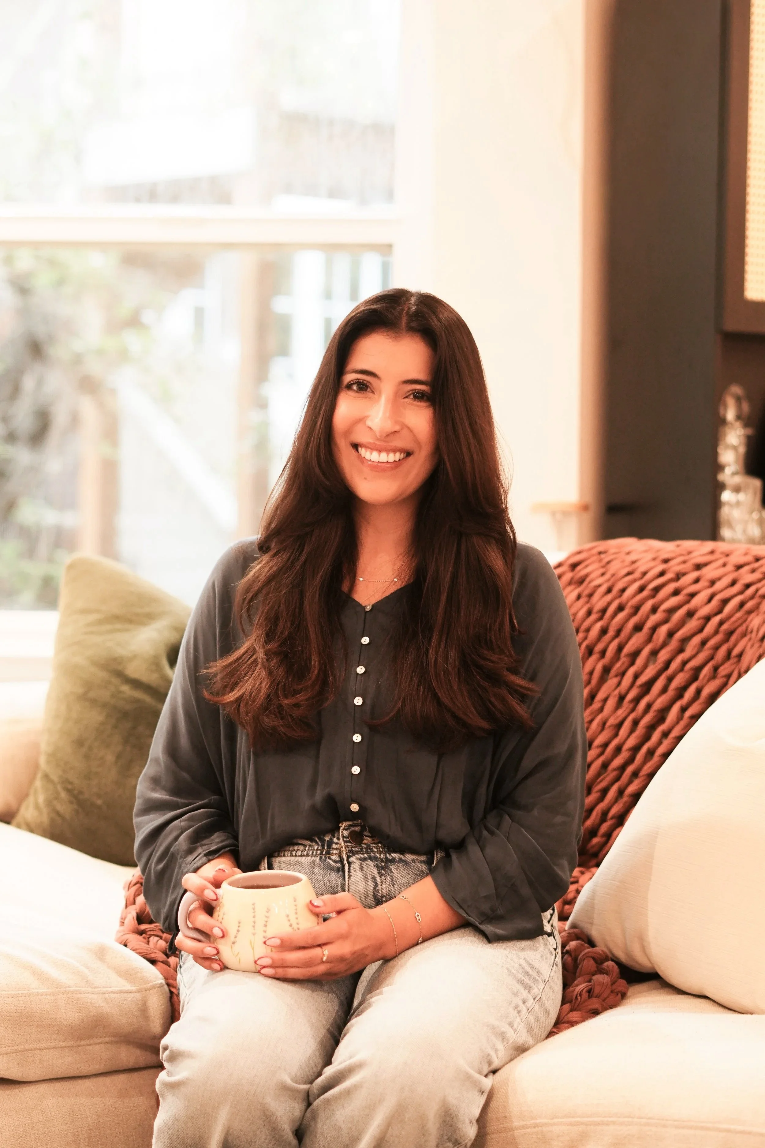 A woman, Nicole, with long dark hair sitting on a sofa, smiling, holding a cup, with a window behind her.