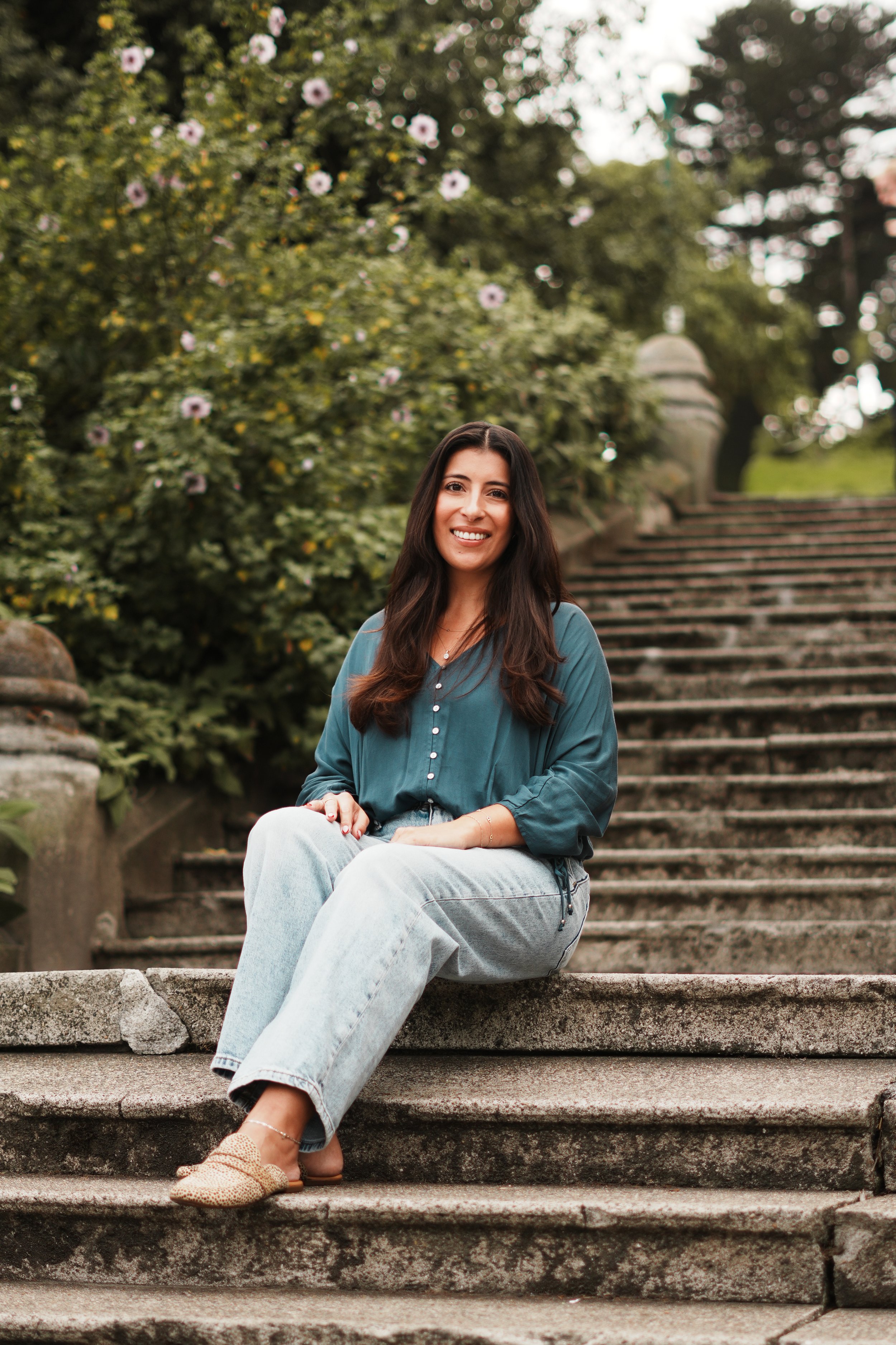 Nicole. A woman with long dark hair, wearing a teal blouse and light jeans, is sitting on stone steps outdoors, smiling at the camera, with greenery and flowers in the background.