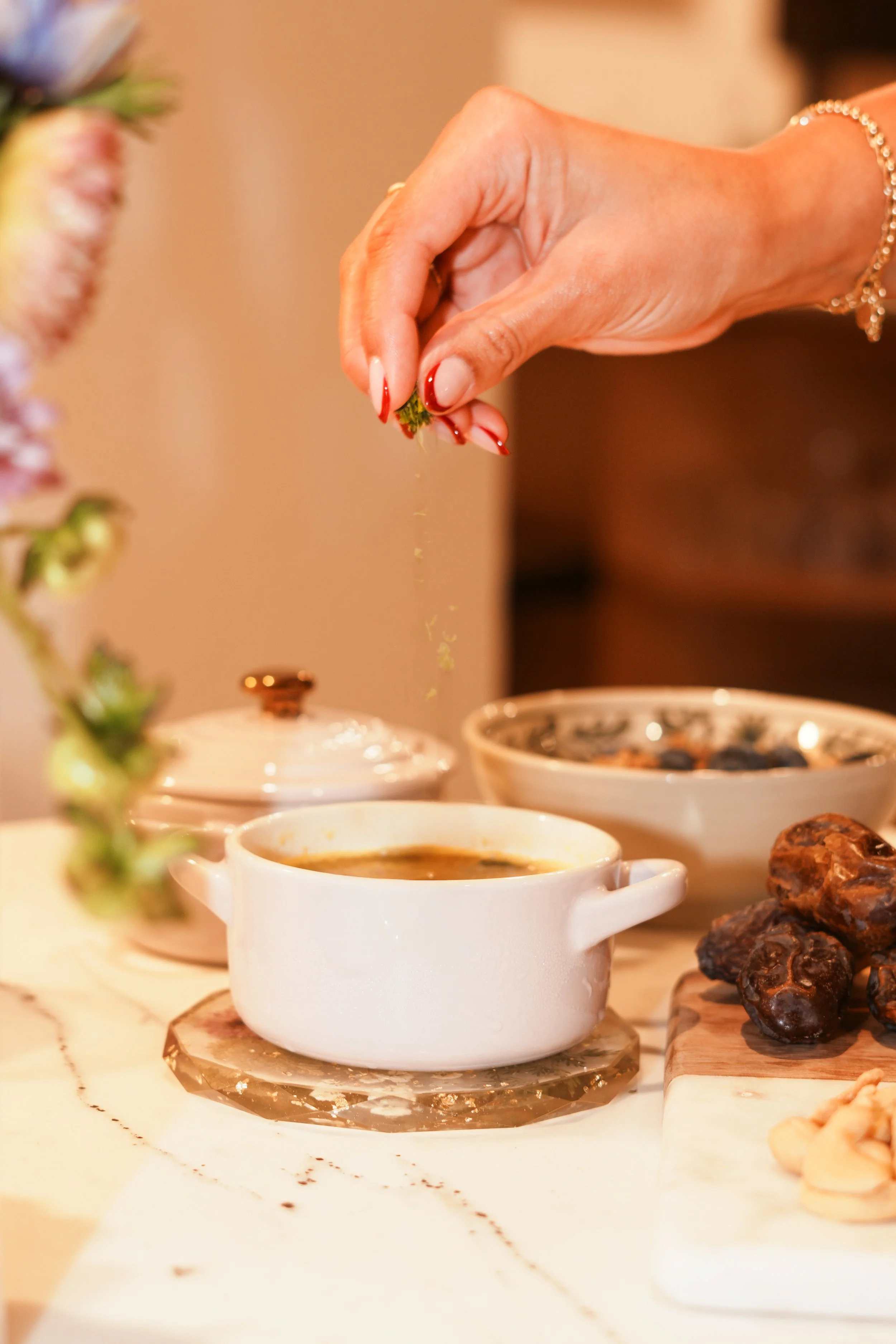 A hand sprinkling green herbs onto a bowl of soup on a marble countertop, with other bowls and dried dates nearby.