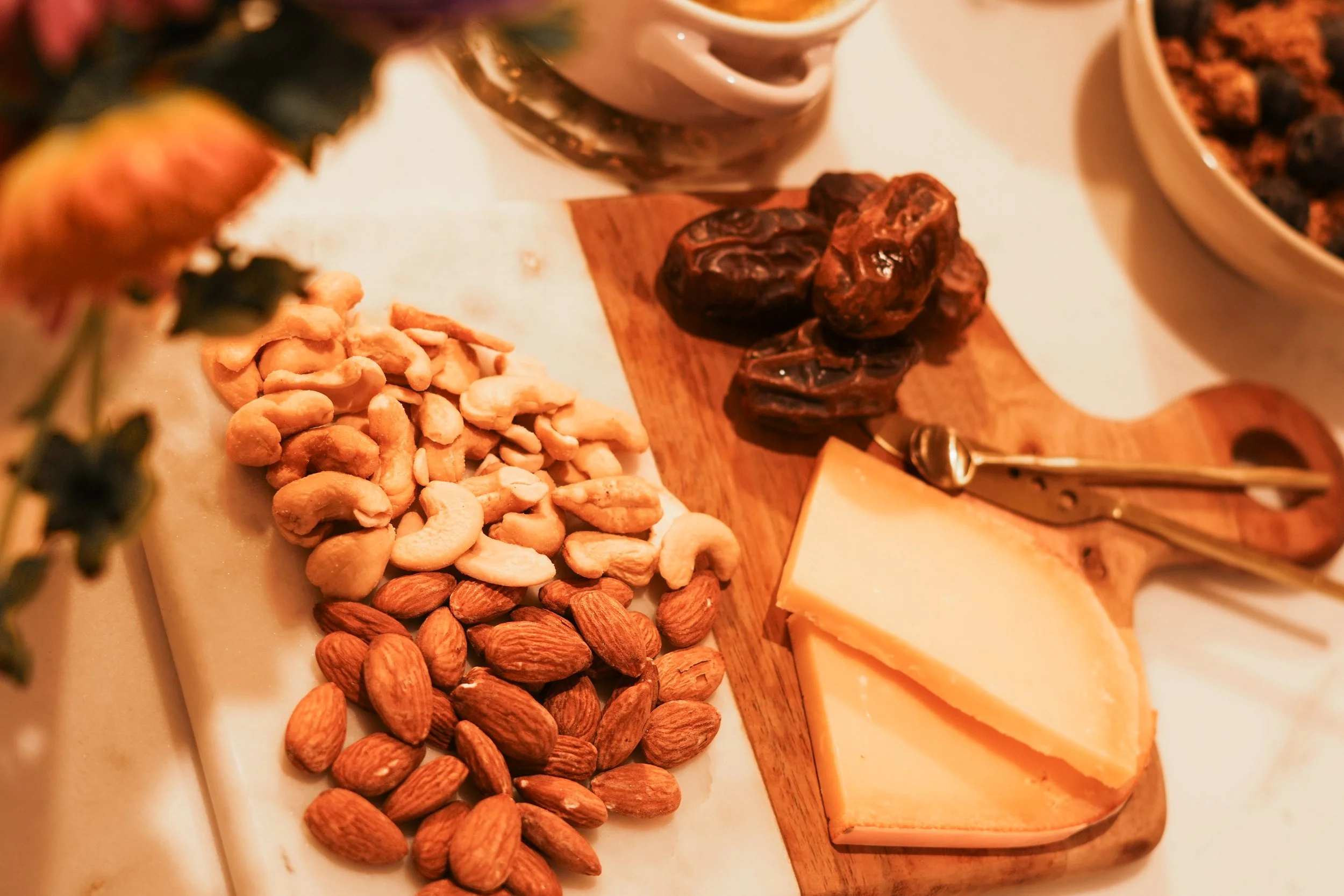 A cheese and nut platter with cheddar cheese, almonds, cashews, and dried dates on a wooden serving board, with bowls of berries and a mug in the background.