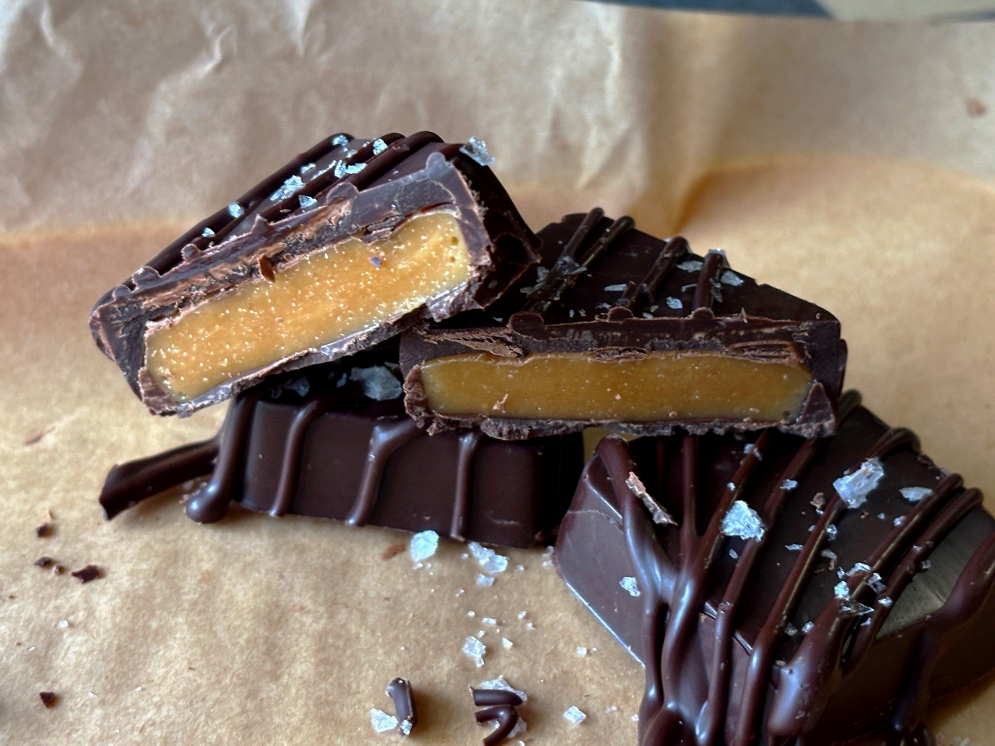 Piece of chocolate candy with a caramel center, decorated with chocolate drizzle and salt flakes, on a parchment paper surface.