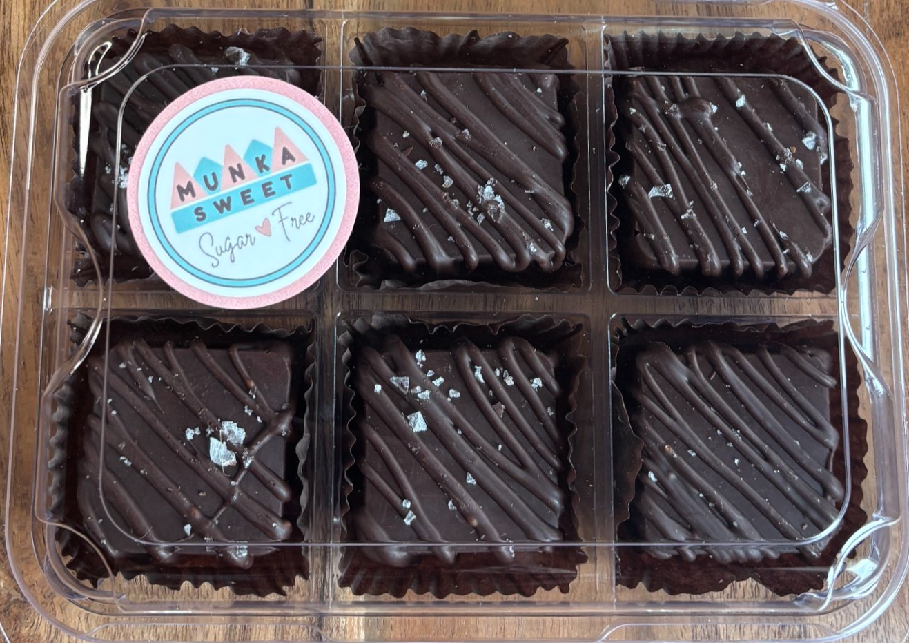 Six pieces of dark chocolate brownie with diagonal chocolate drizzle and sea salt flakes in a clear plastic container with a circular label that reads "MUNKA SWEET, Sugar Free," on a wooden surface.