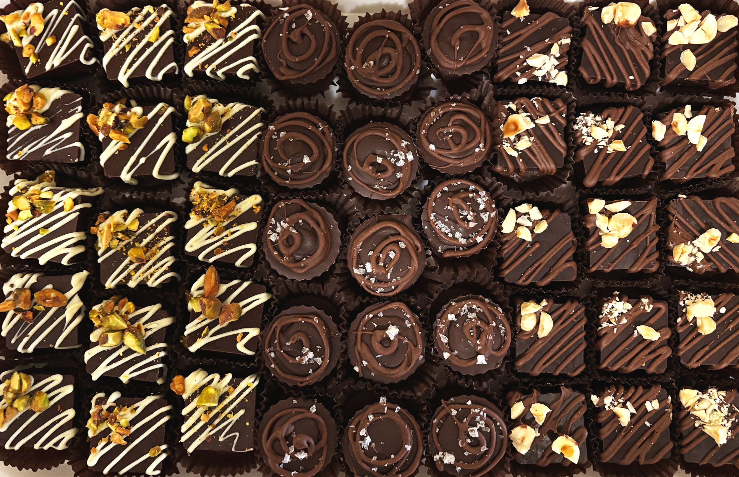 A variety of assorted chocolates and bonbons decorated with white and dark chocolate drizzle, chopped nuts, and salt flakes.