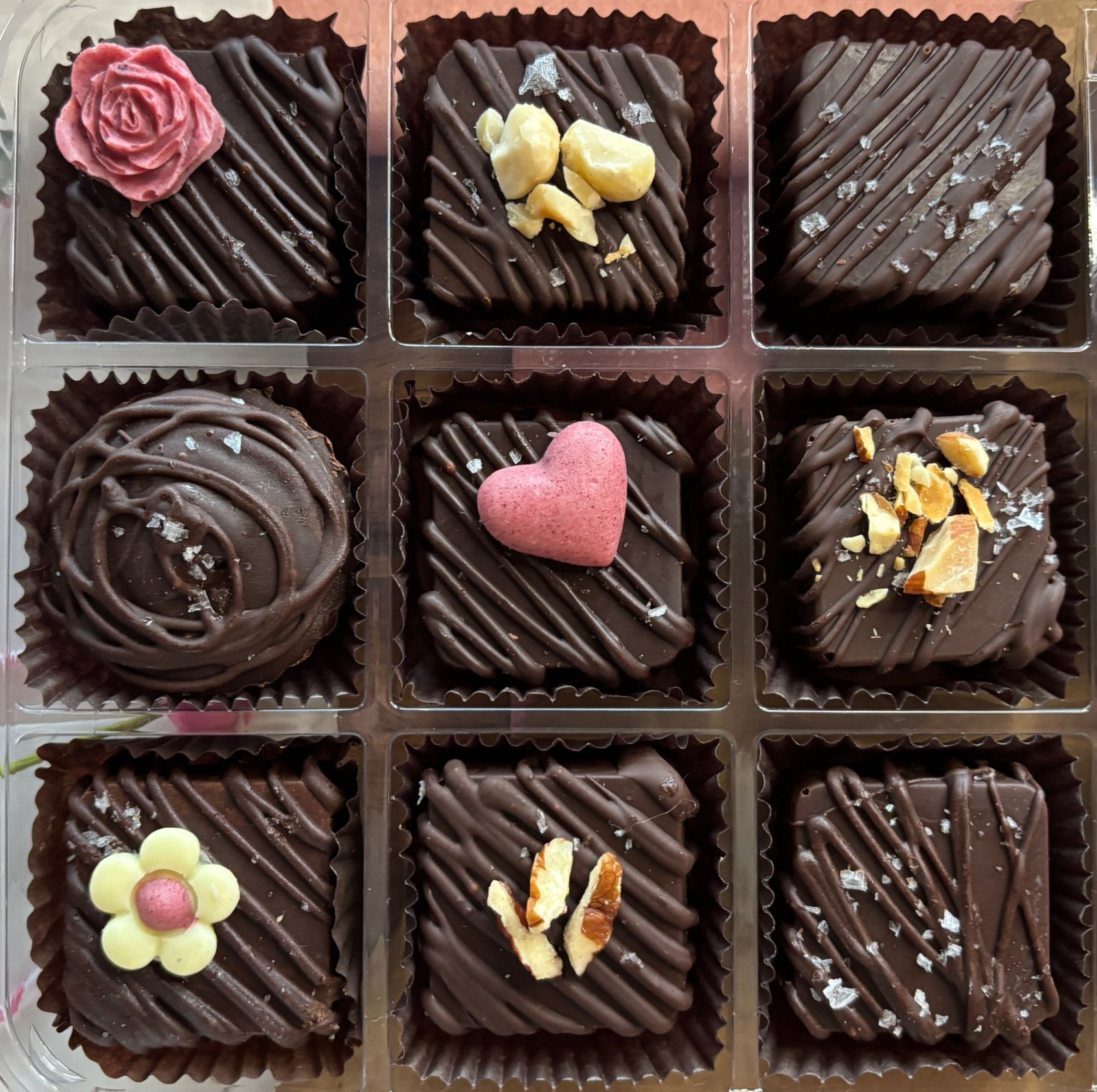 A box of assorted chocolates with various decorations including pink roses, yellow and pink hearts, a flower, and crunchy nuts on top.