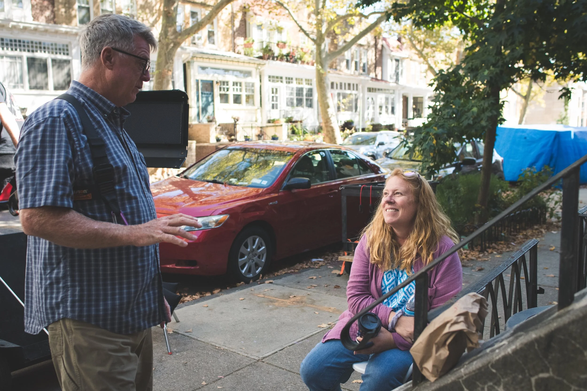 A man and a woman having a conversation on a sidewalk in a residential neighborhood. The woman is sitting on a bench, smiling, with a drink and a paper bag beside her. The man is standing, holding a mobile phone. In the background, there are parked c