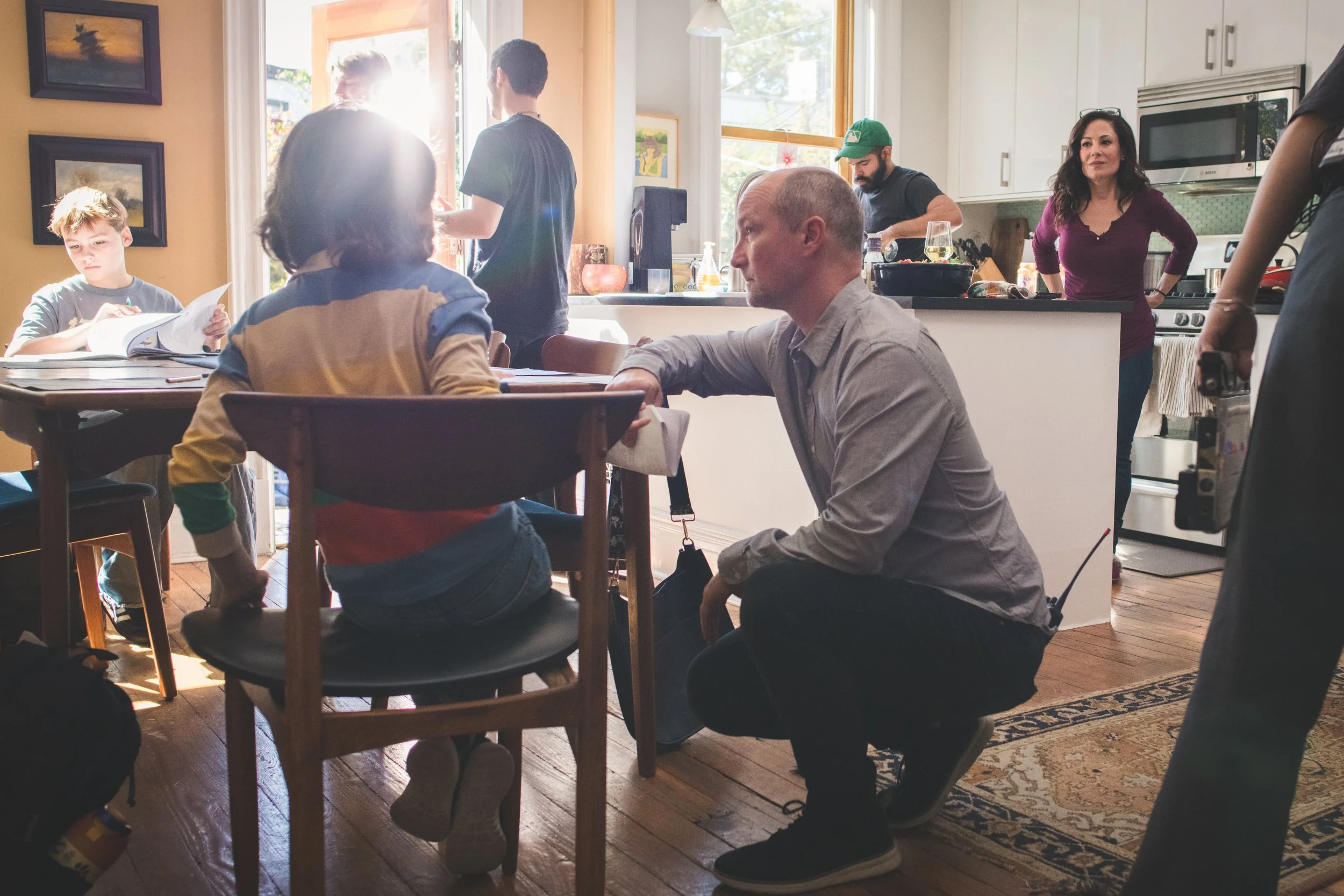 Family gathered in the kitchen and dining area of their home, with a man kneeling beside a young girl sitting at a dining table, a woman standing and looking at others in the kitchen, and a boy reading a book at the table. Other family members are pr