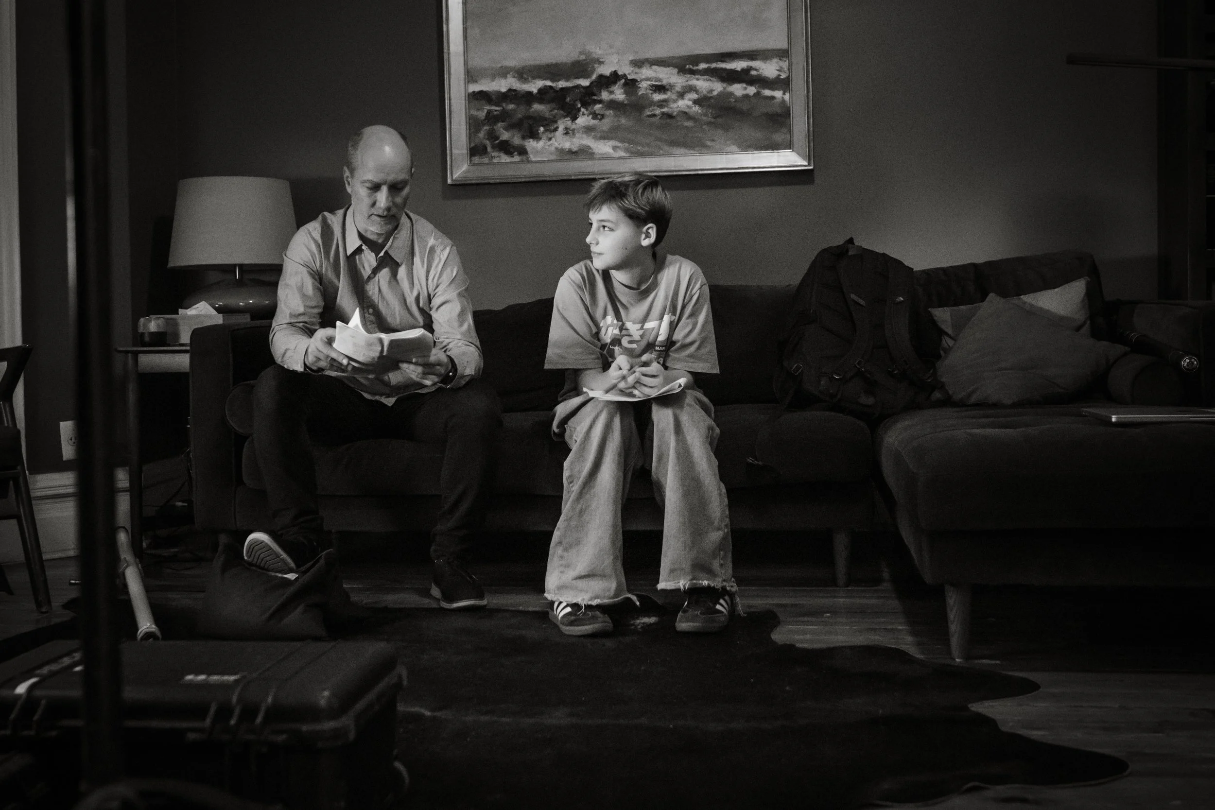 An older man and a young boy sitting on a dark sofa in a living room. The man is reading a book, and the boy is holding a notebook, looking at the man. There is a painting on the wall behind them, a lamp on a side table, and backpacks and pillows on 