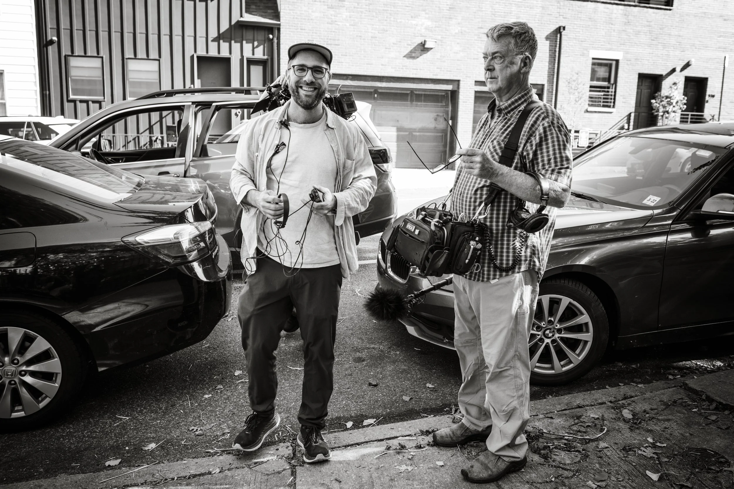 Two men standing next to parked cars on a city street, talking and holding recording equipment, one smiling and the other looking serious.