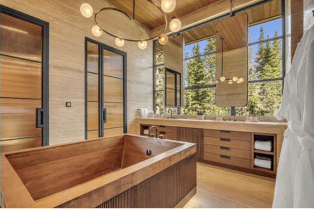 Modern bathroom with a wooden bathtub, large windows showing outdoor trees, black-framed glass closet doors, and contemporary lighting fixtures.