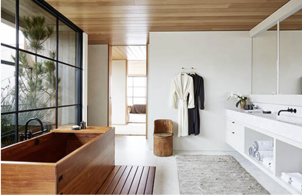 Modern bathroom with wooden soaking tub, large window with view of plants, white walls, and built-in white cabinet.