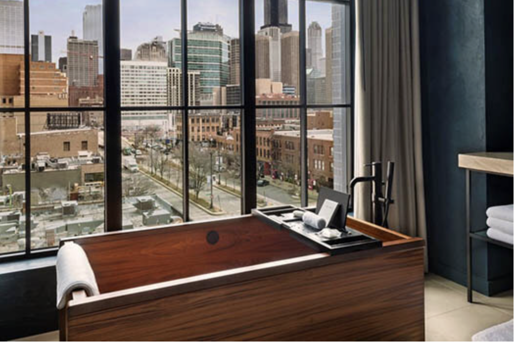 A modern hotel room with a wooden bathtub near a large window showing a cityscape with tall buildings.