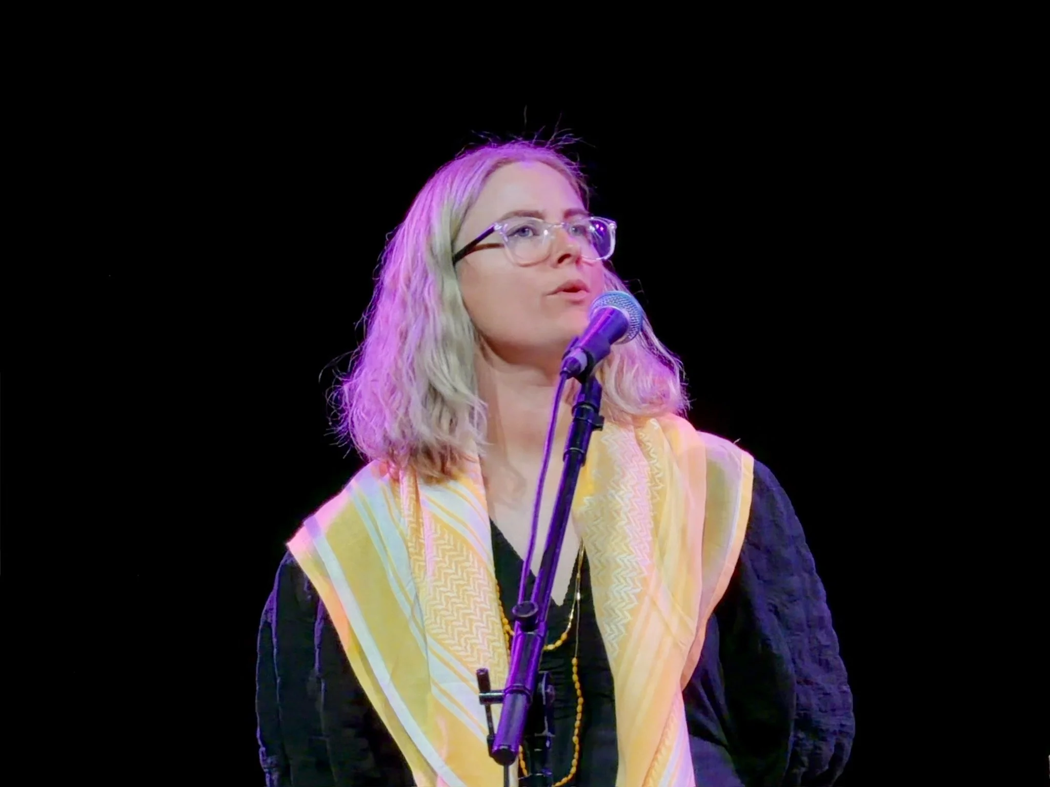 A woman with shoulder-length blonde hair and glasses, wearing a yellow scarf, speaking into a microphone on stage against a black background.