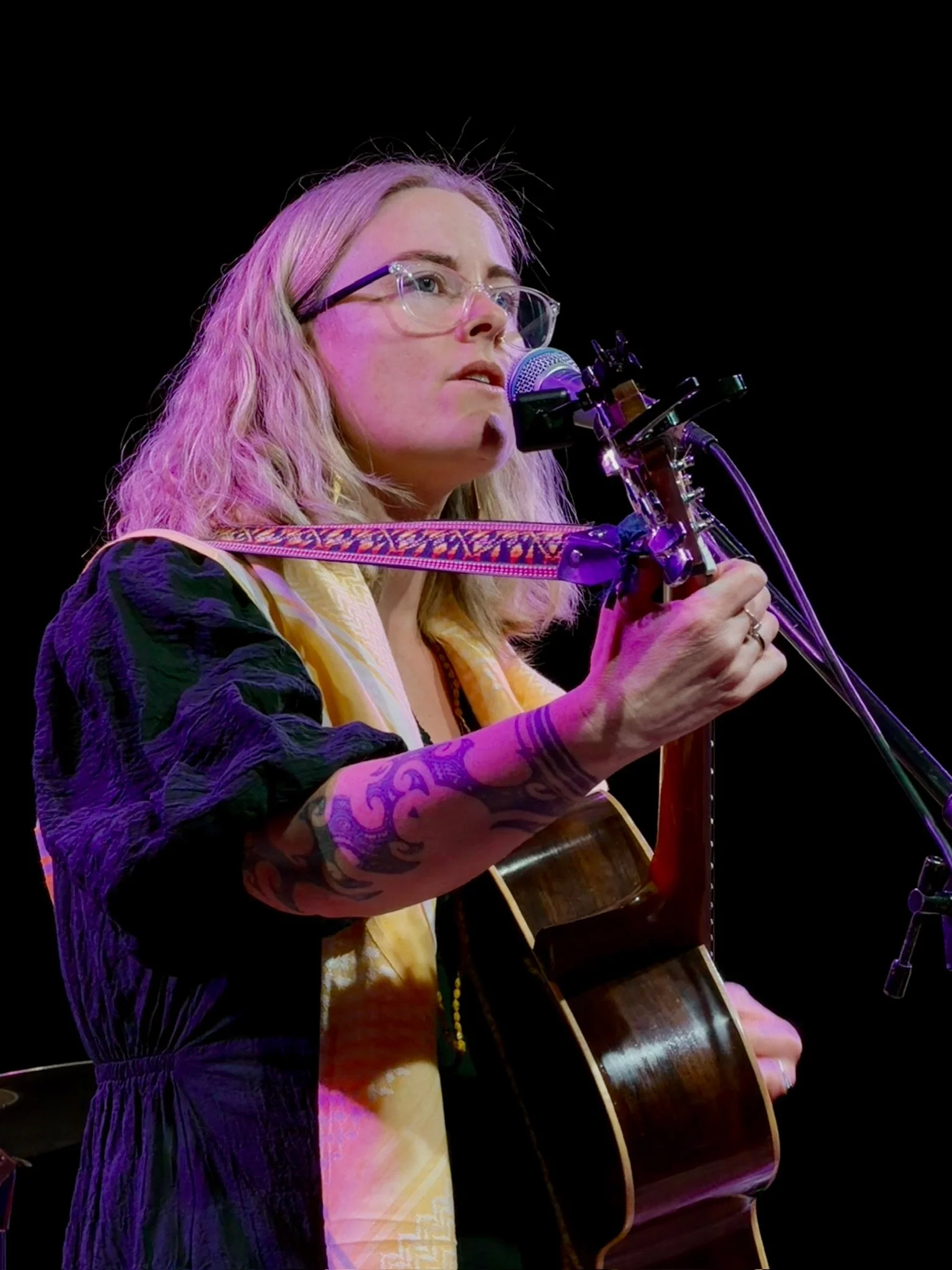 Female musician with blonde wavy hair, glasses, and tattoos playing an acoustic guitar and singing into a microphone on stage against a black background.