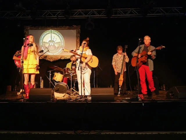 A band performing on stage with five members, including a drummer, two guitarists, a keyboardist, and a female vocalist. The background features a logo of the Whitlington Folk Arts Council.