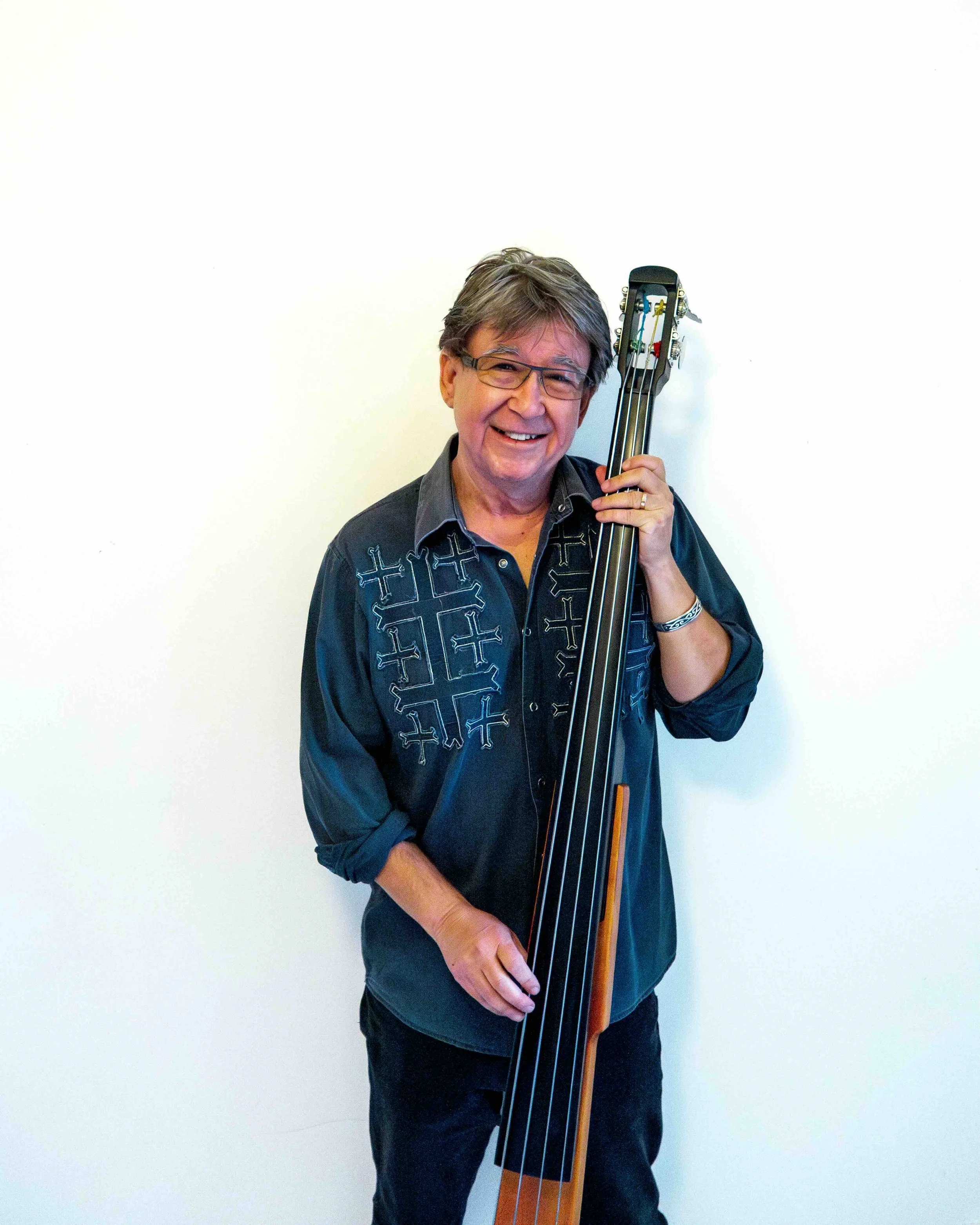 A man with gray hair and glasses smiling, holding a large upright bass, standing against a plain white wall.