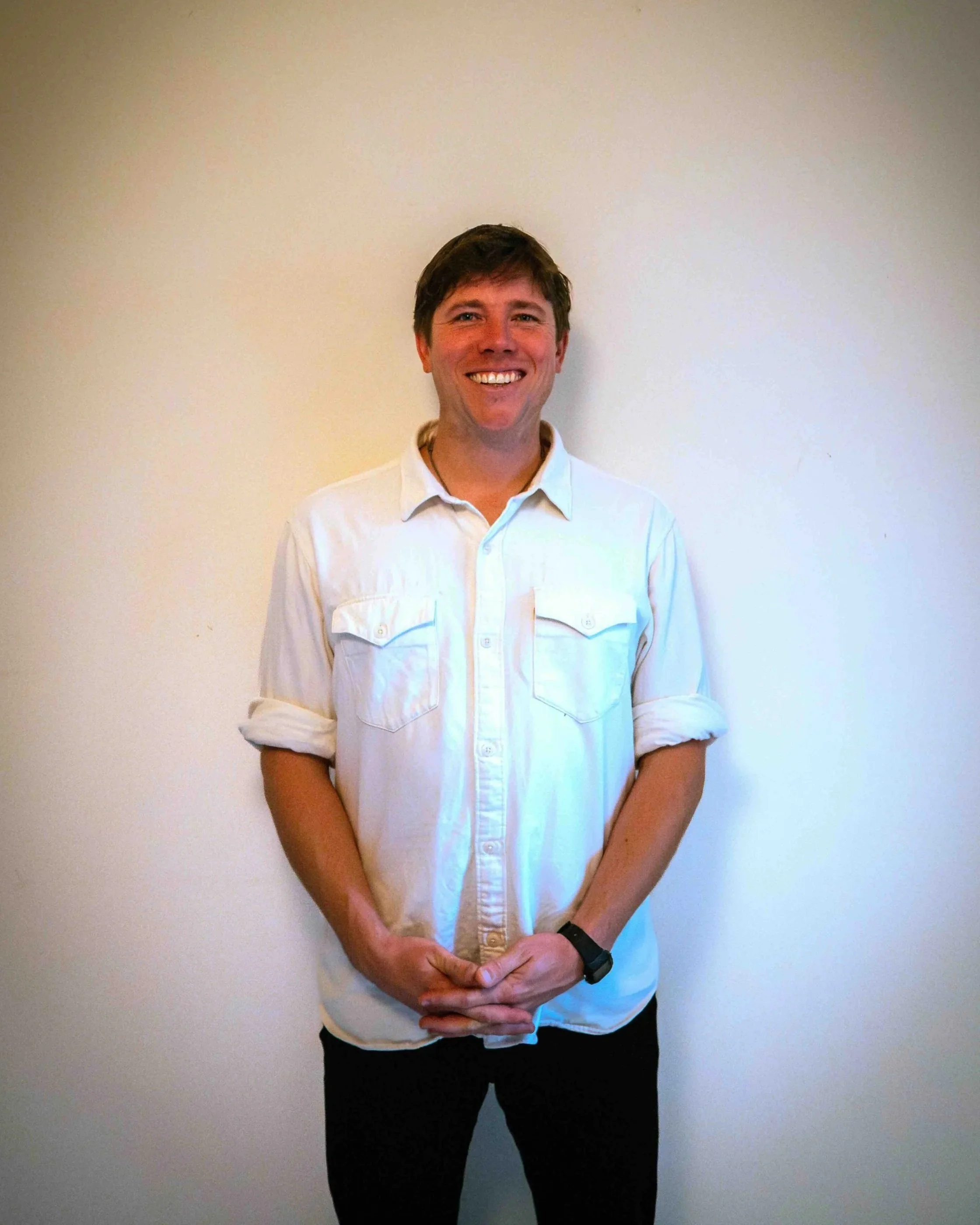 A smiling man with short brown hair wearing a white button-up shirt with rolled-up sleeves, standing against a plain off-white wall.