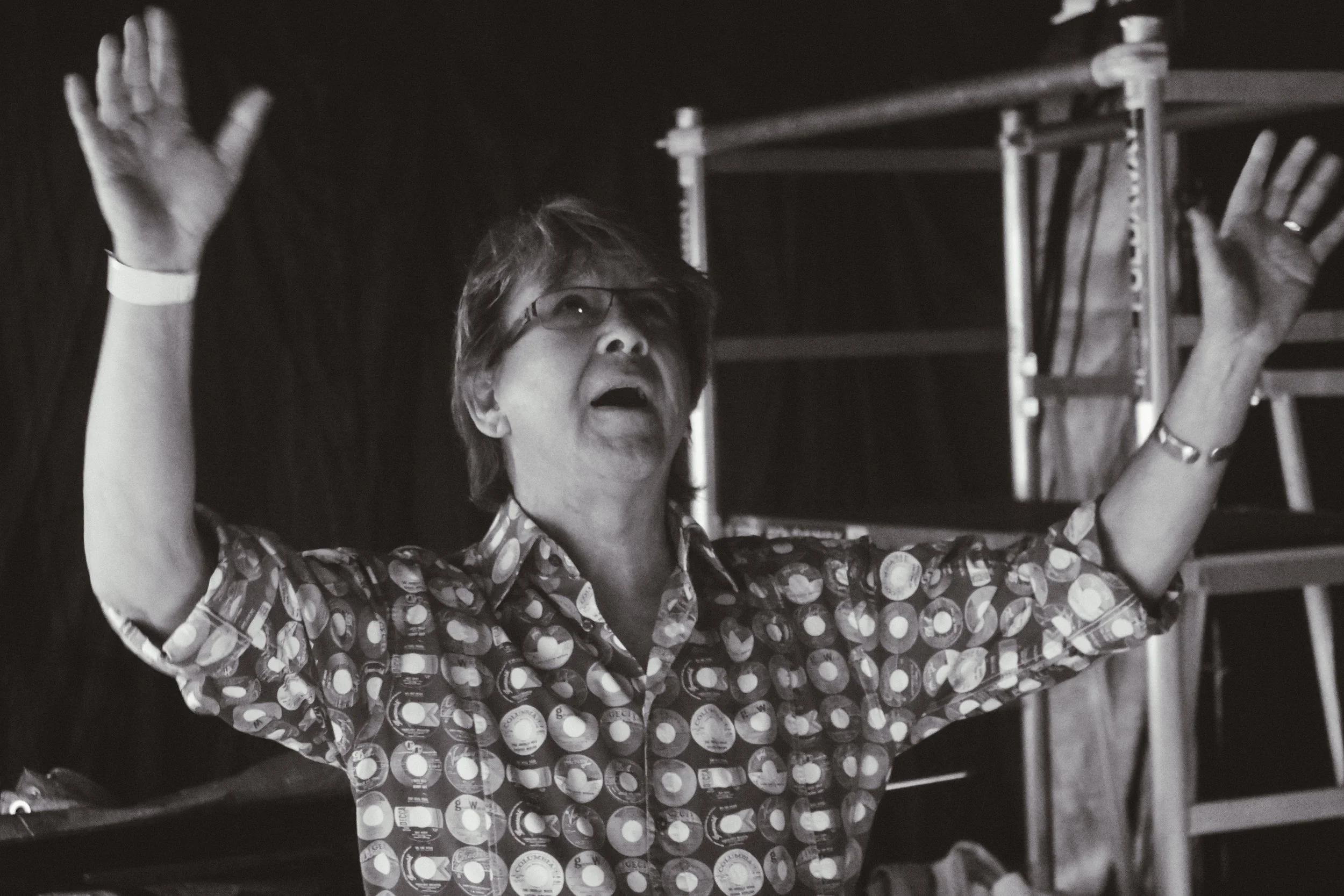 A woman with glasses, short hair, wearing a fantasy print shirt, raising her arms with her eyes closed and mouth open, standing in front of a black curtain and a metal shelf.