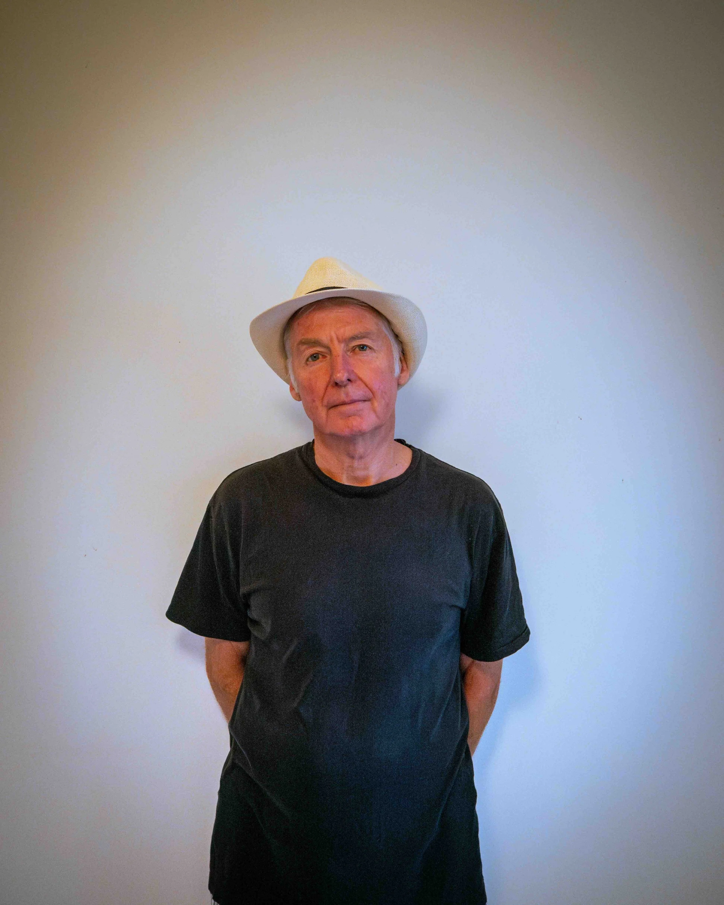 Older man with gray hair wearing a black T-shirt and a white hat, standing against a plain white wall.