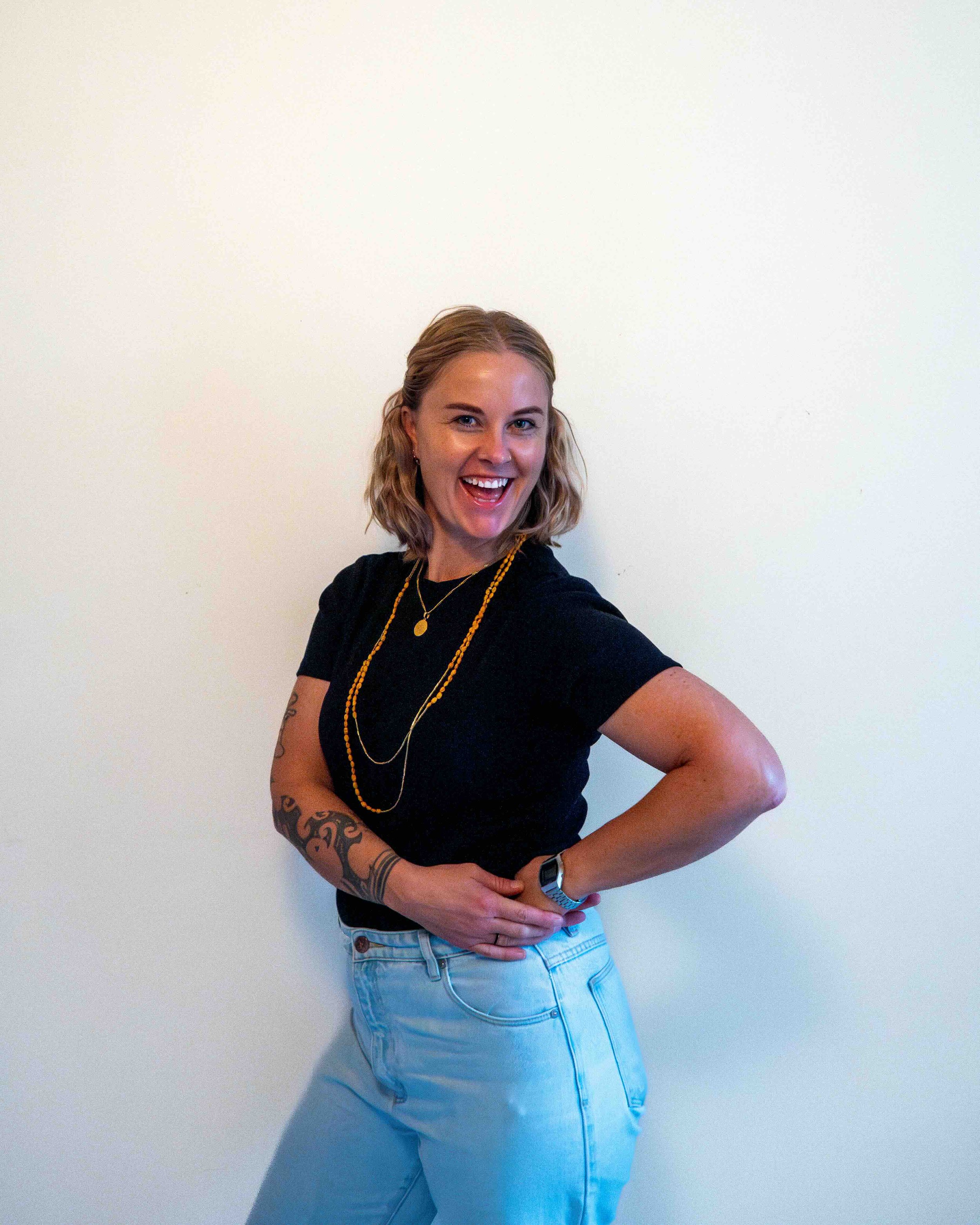 A woman with blonde hair smiling, wearing a black t-shirt, light blue jeans, and layered necklaces, posing against a plain light-colored wall.