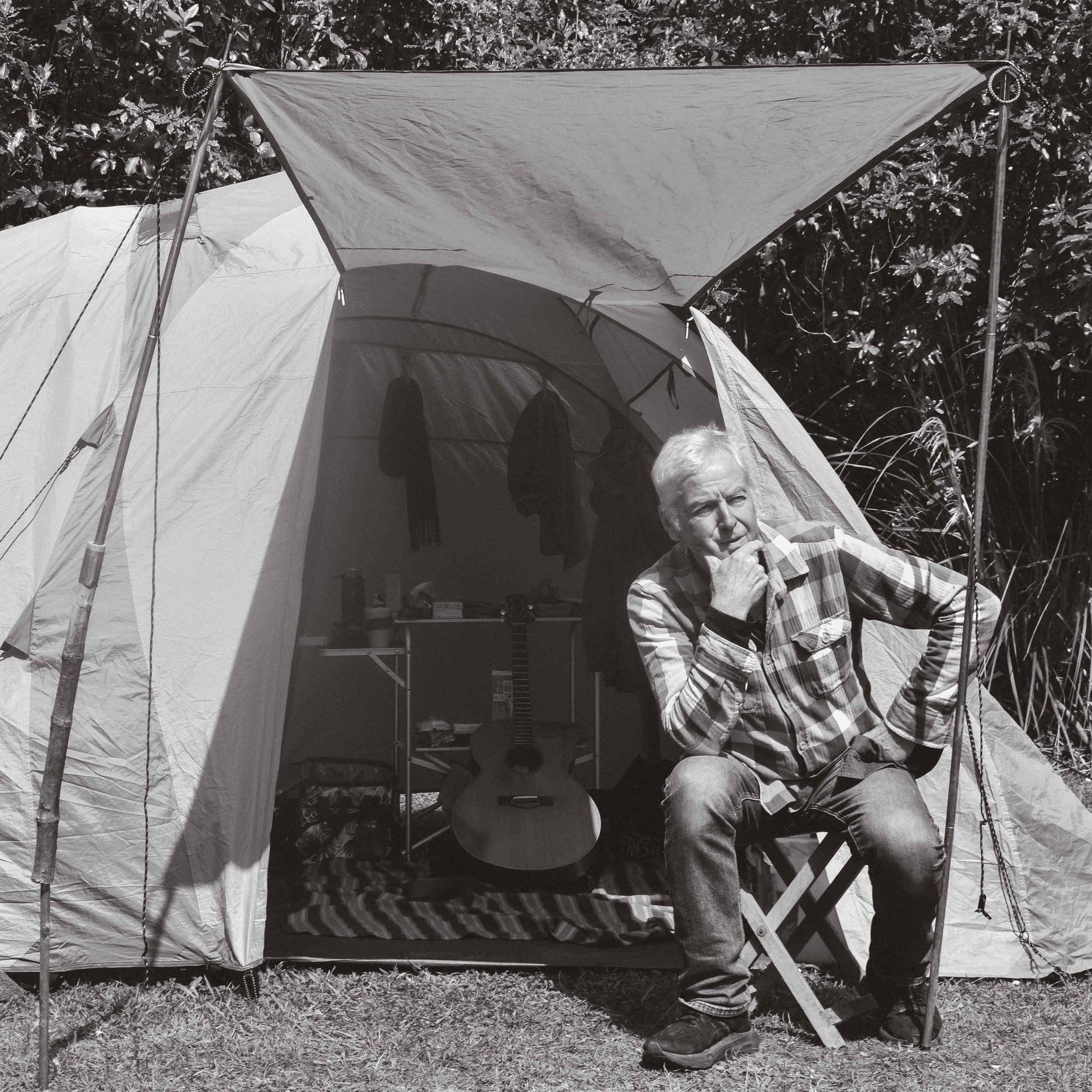 An older man sits outside a camping tent with a thoughtful expression, wearing a plaid shirt and jeans. Inside the tent, there is an acoustic guitar, some clothing hanging, and a small table with items on it. The scene appears to be in a grassy outdoor area surrounded by trees.
