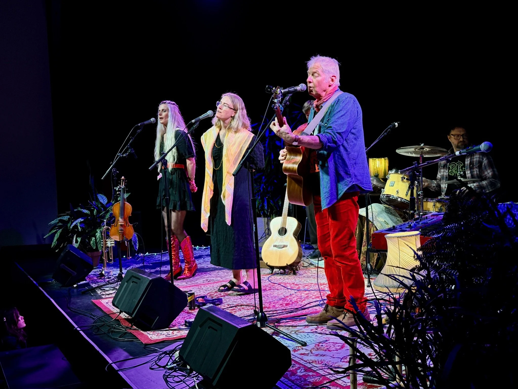 A musical band performing live on stage with three women singing into microphones, a man playing guitar, and a drummer playing drums in the background. The stage has plants and a carpet.