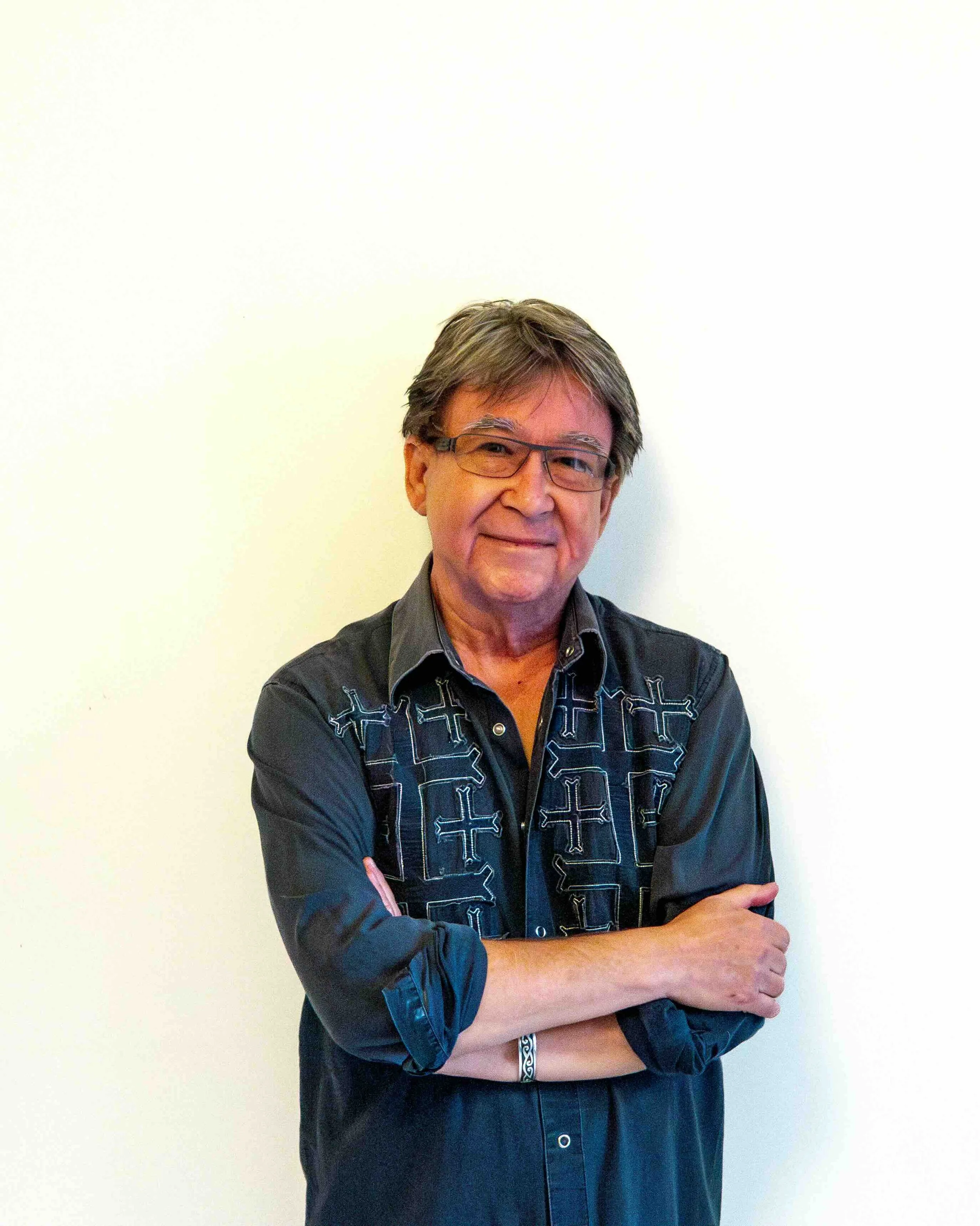 A man wearing glasses and a black shirt standing against a plain light-colored wall.