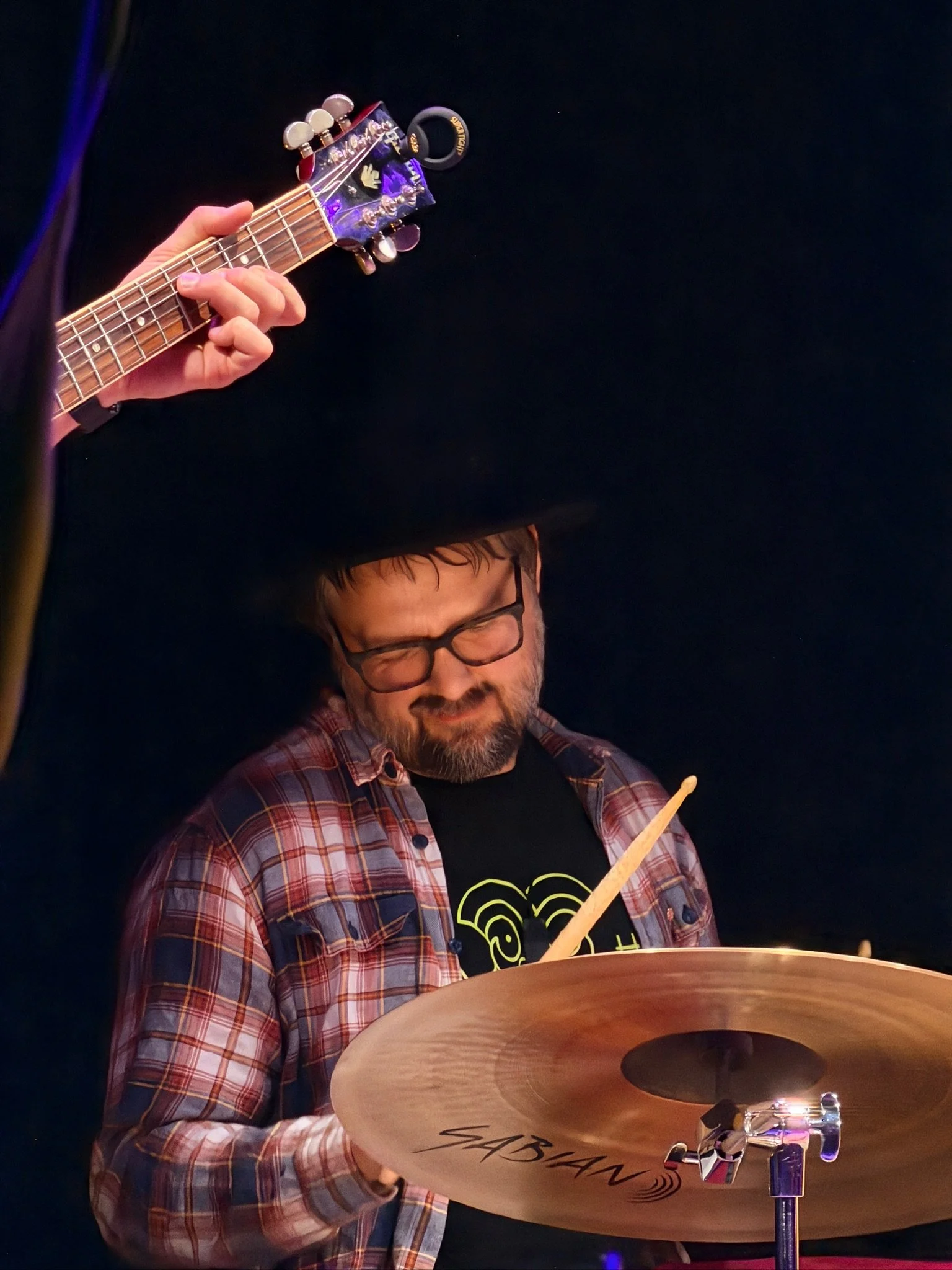 A man with glasses and a beard is playing a cymbal, with a person playing an electric guitar above him. The setting appears to be a dark indoor space, possibly a stage.
