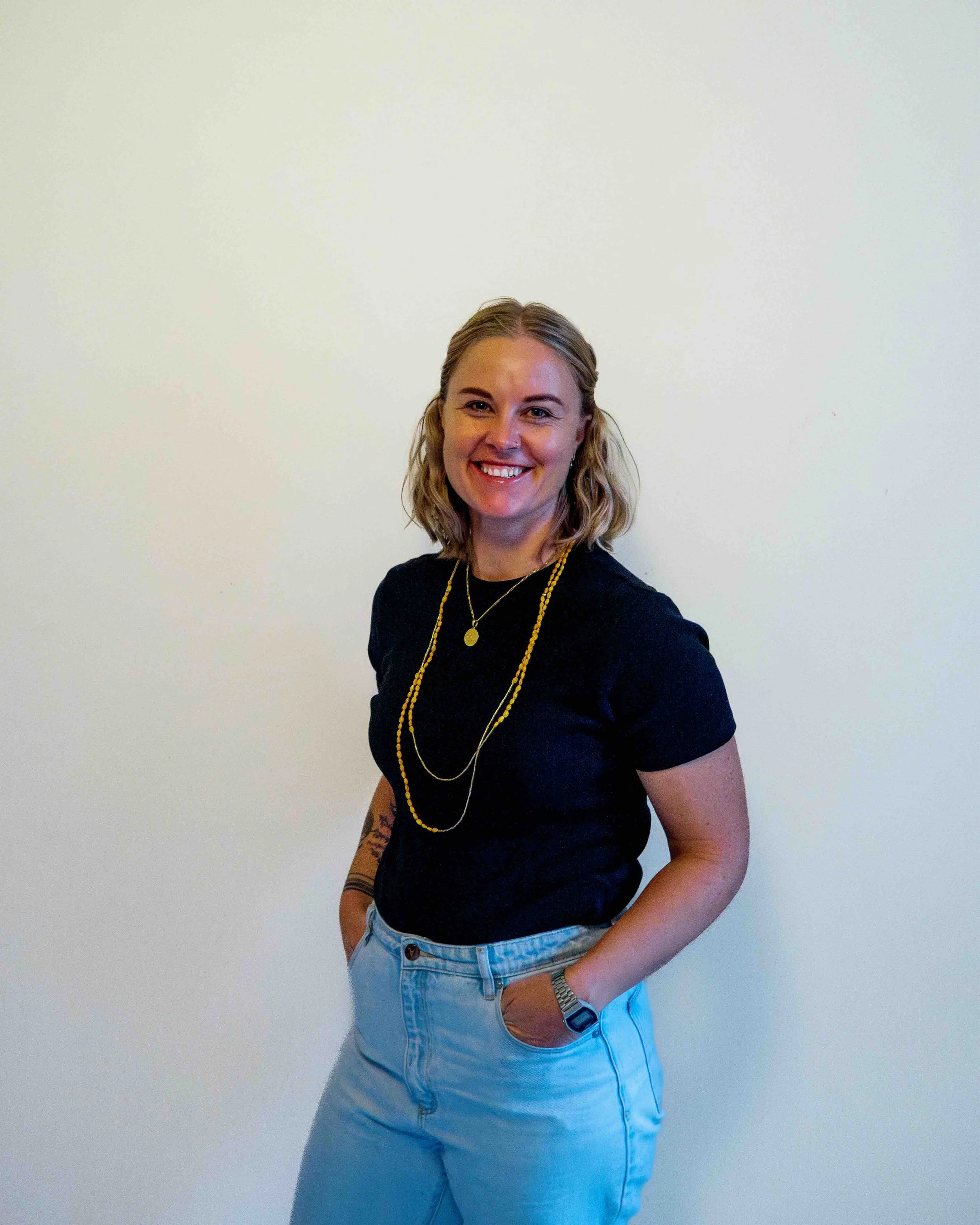 A woman with blonde wavy hair smiling, wearing a black t-shirt, light blue jeans, layered gold necklaces, with her hands in her pockets against a plain white wall.