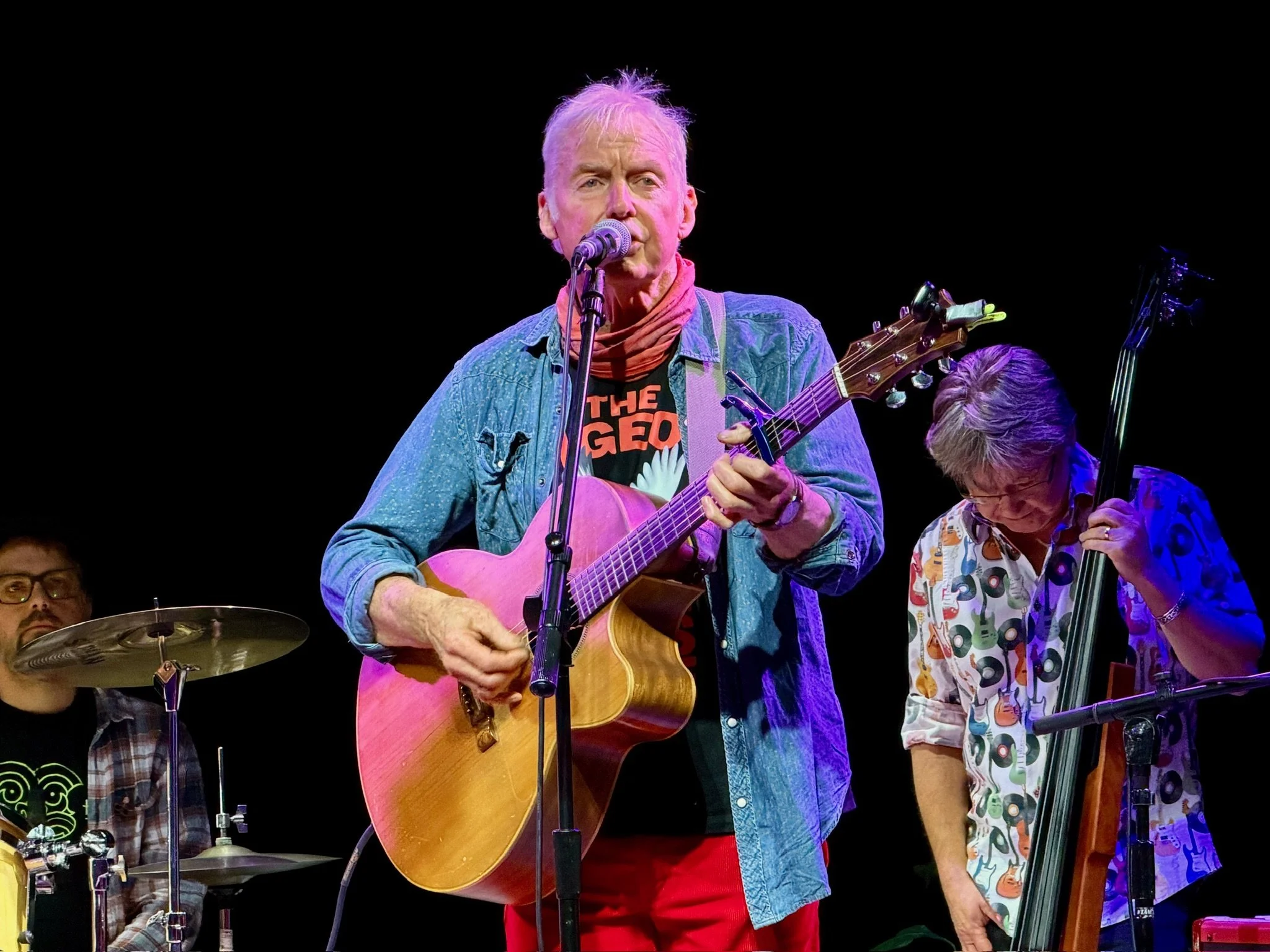 A band performing on stage with a man singing and playing guitar, a woman playing bass, and a person playing drums in the background.