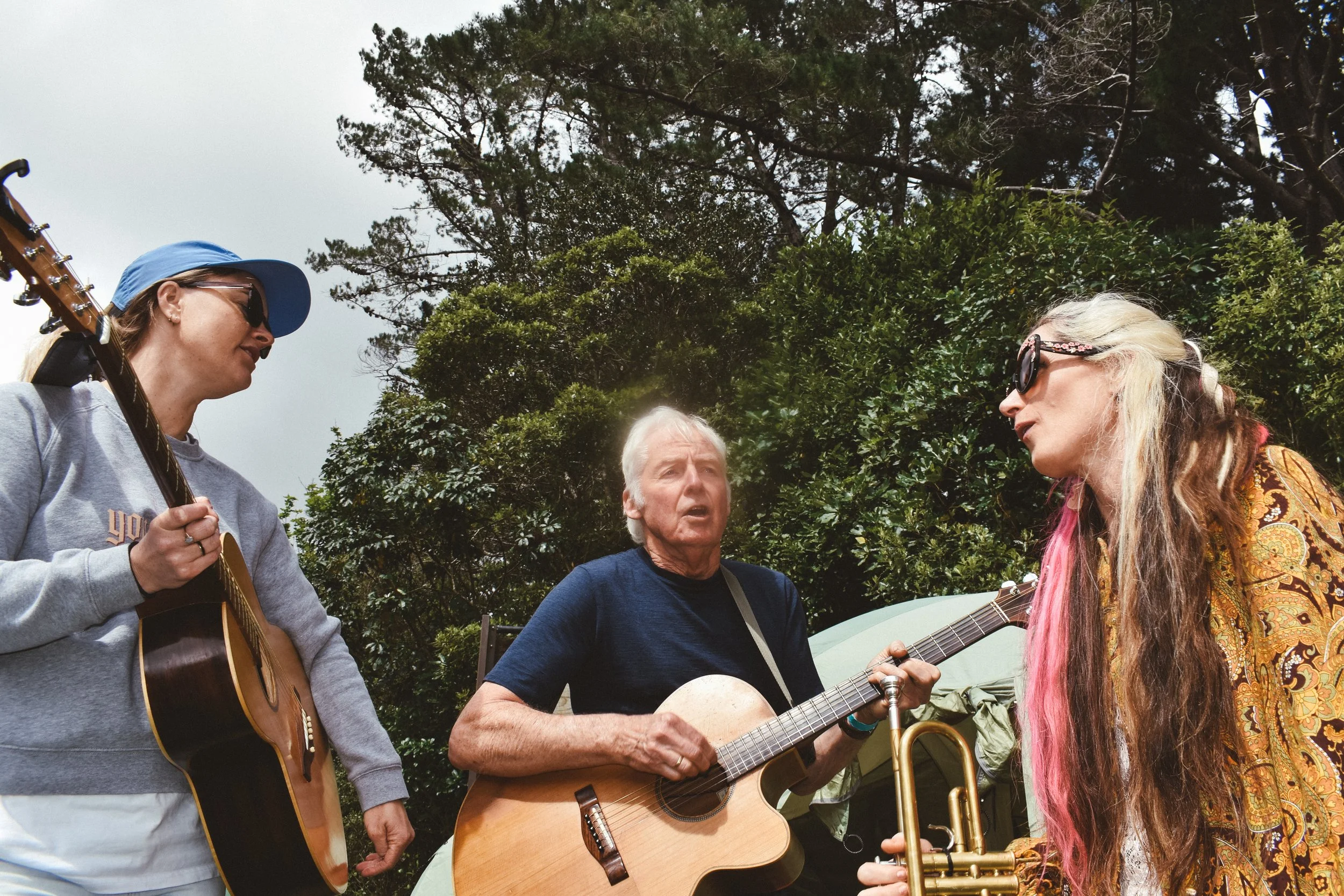 Three musicians outdoors, two women playing guitars and a woman holding a trumpet, beside trees and a tent.