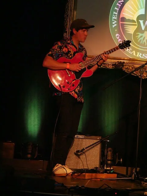 Young man performing on stage playing an electric guitar, wearing a colorful floral shirt, black pants, white sneakers, and a black cap, with a logo and celebration text projected on the background.