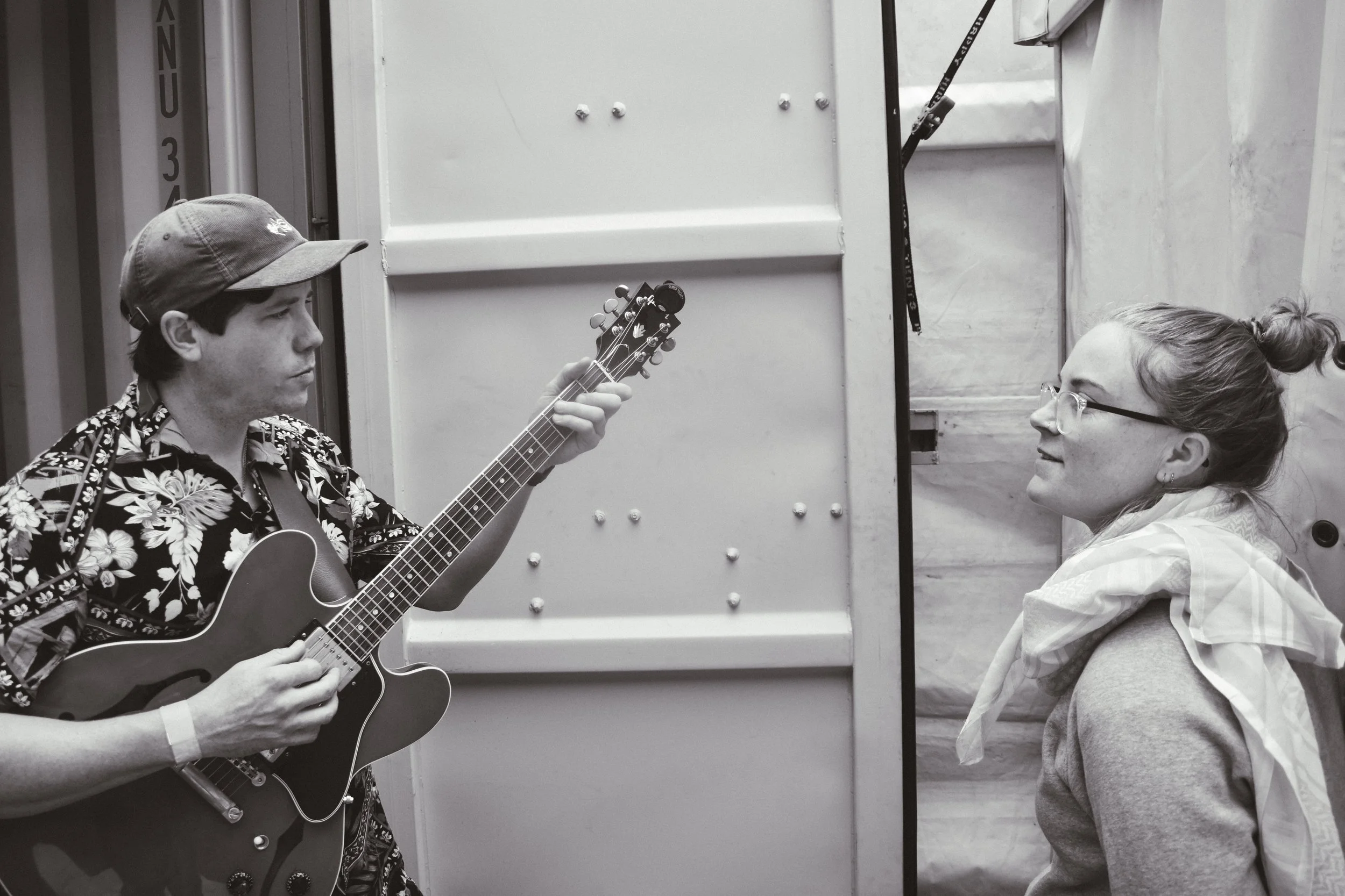 A young man wearing a hat and a Hawaiian shirt playing an electric guitar, facing a young woman with glasses and a hoodie, who is looking and smiling at him.