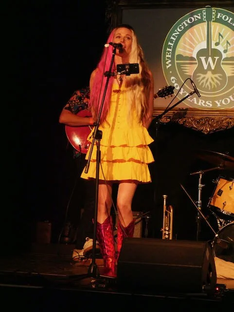 Female singer in a yellow dress and red boots performing on stage with a microphone, in front of a 