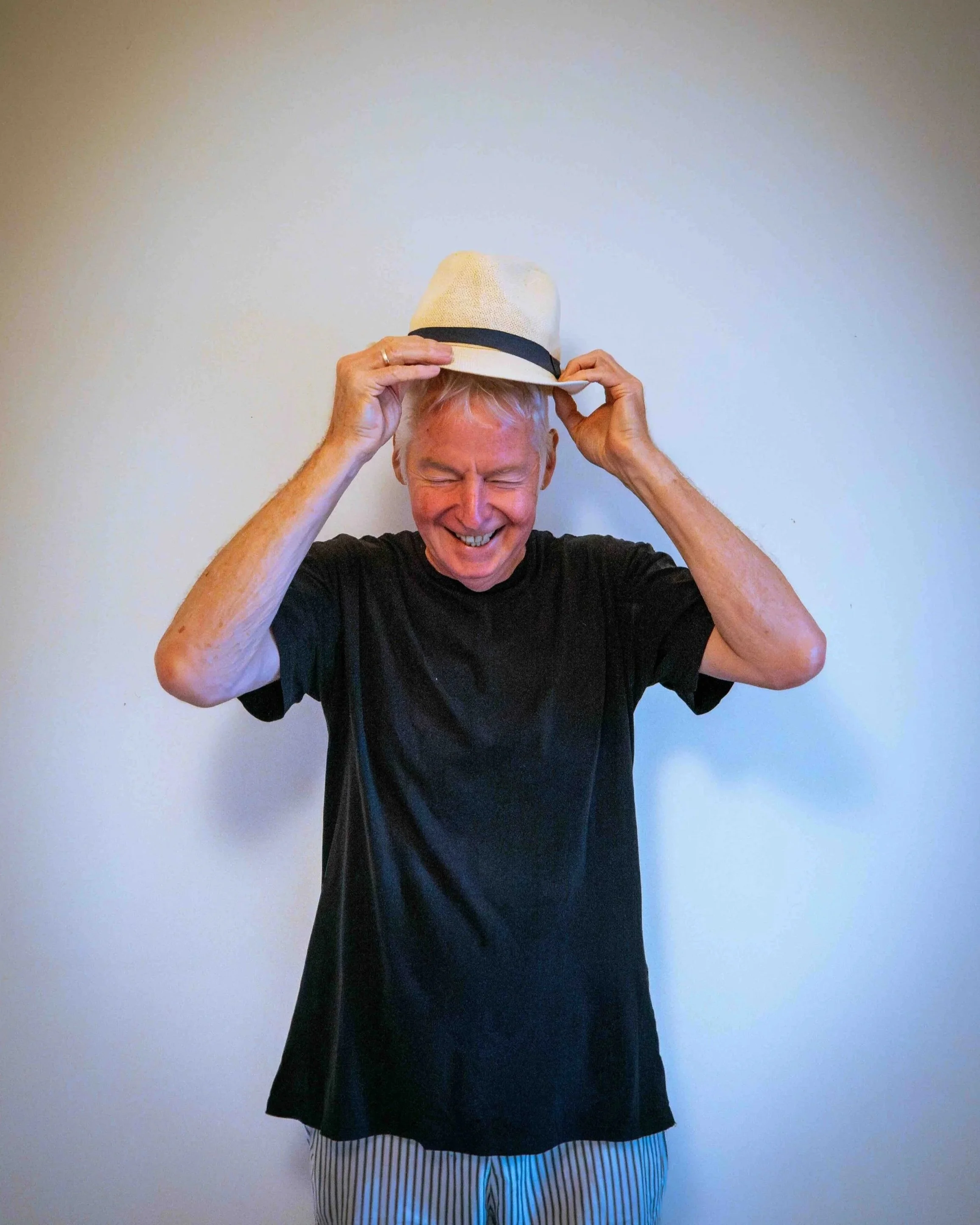 A cheerful man with gray hair, wearing a black t-shirt and striped pants, placing a white fedora hat on his head against a plain light-colored wall.