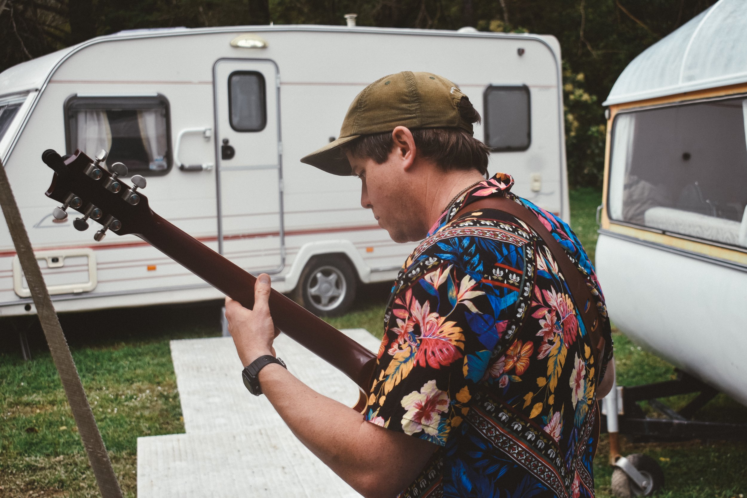 A man playing an acoustic guitar outdoors near a camping trailer and a camper van.