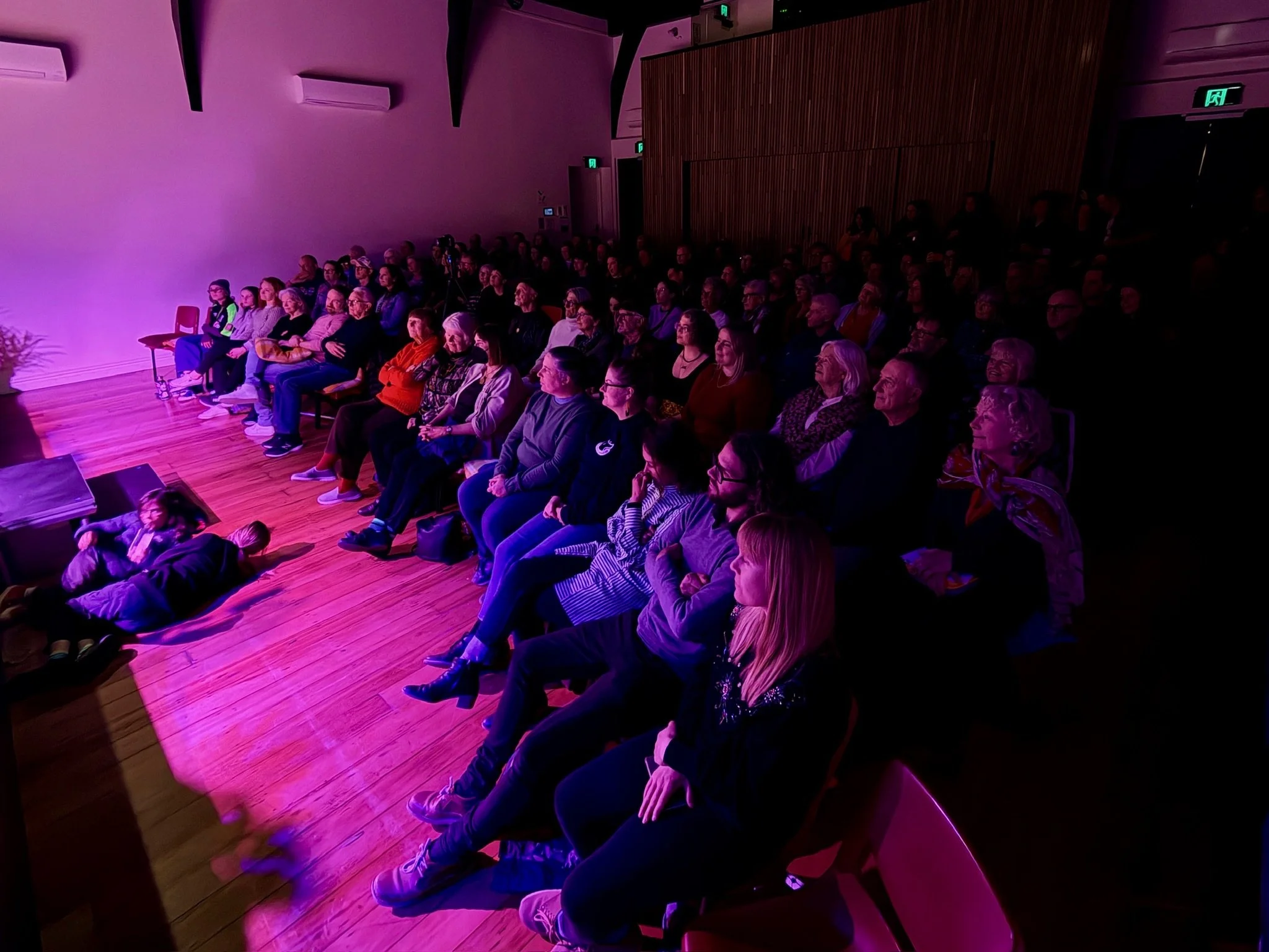 Audience watching a performance or presentation in a dimly lit indoor auditorium with purple lighting.