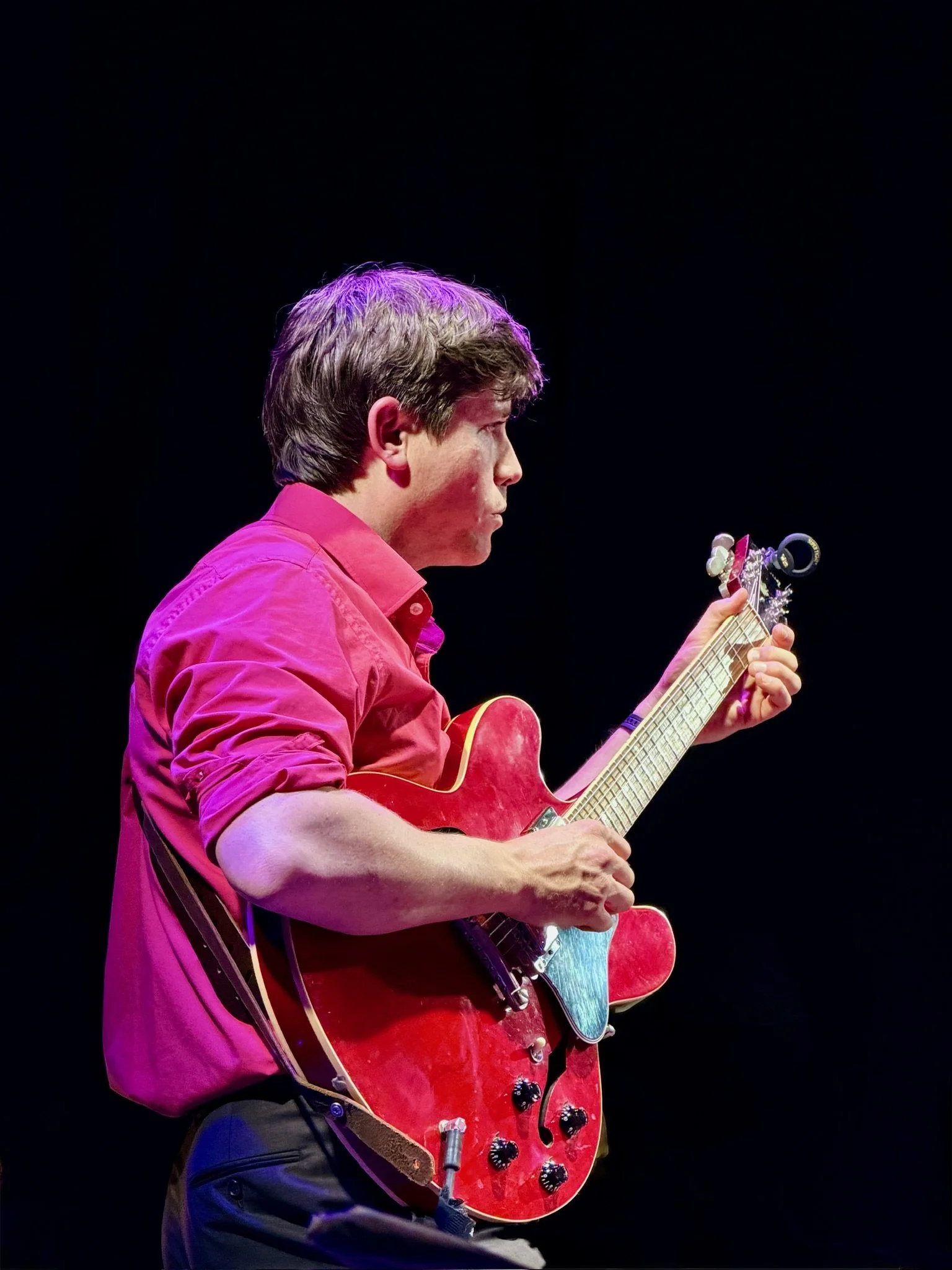 Man playing a red electric guitar on stage with a black background, wearing a pink shirt.