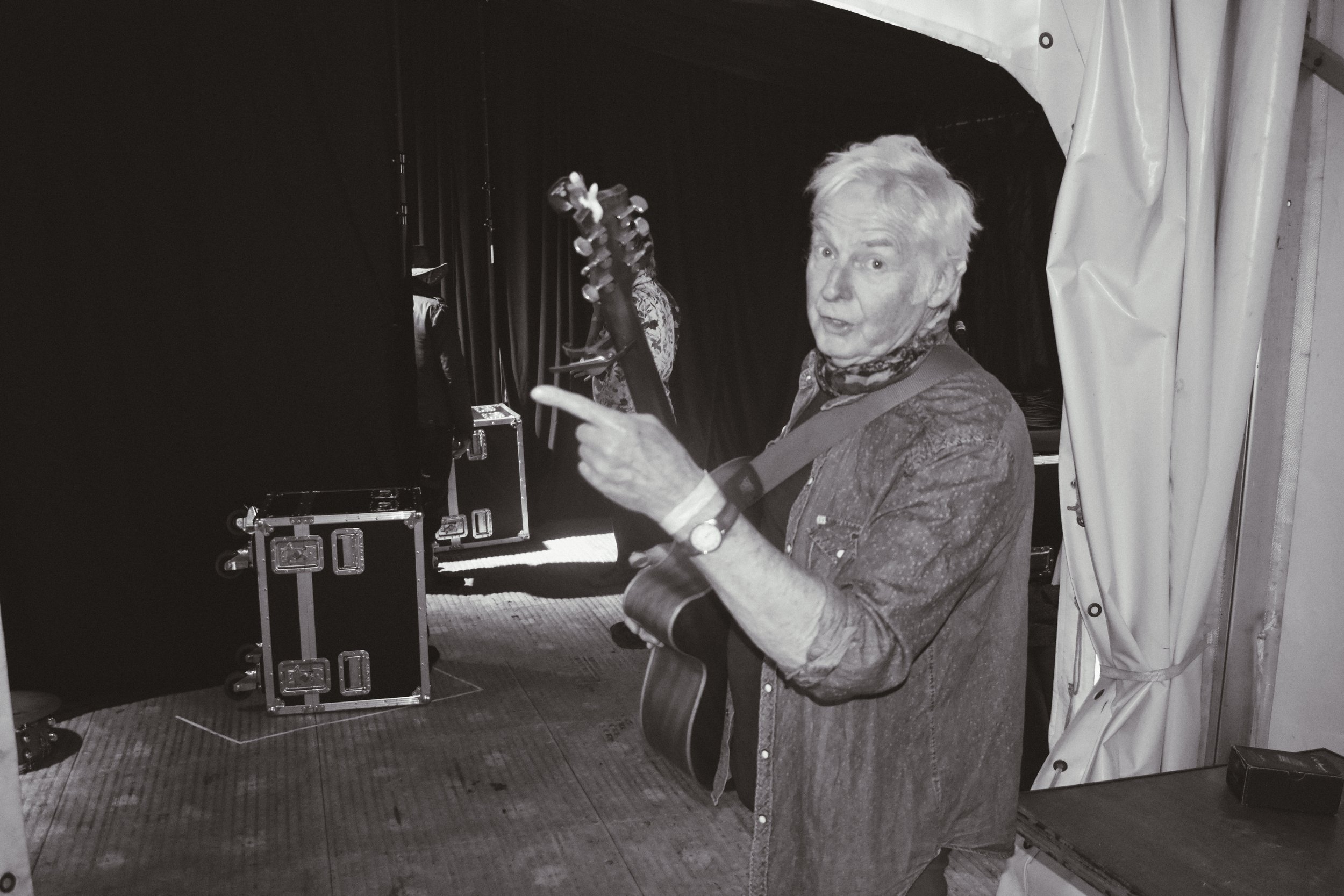 An elderly man with white hair, wearing a watch, a patterned scarf, and a denim jacket, stands backstage with a guitar, pointing and looking toward the camera, with stage equipment and curtains in the background.
