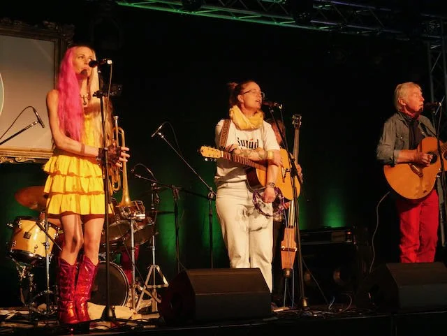 Three musicians performing on stage with microphones, guitar, and drums in the background, under green stage lighting.