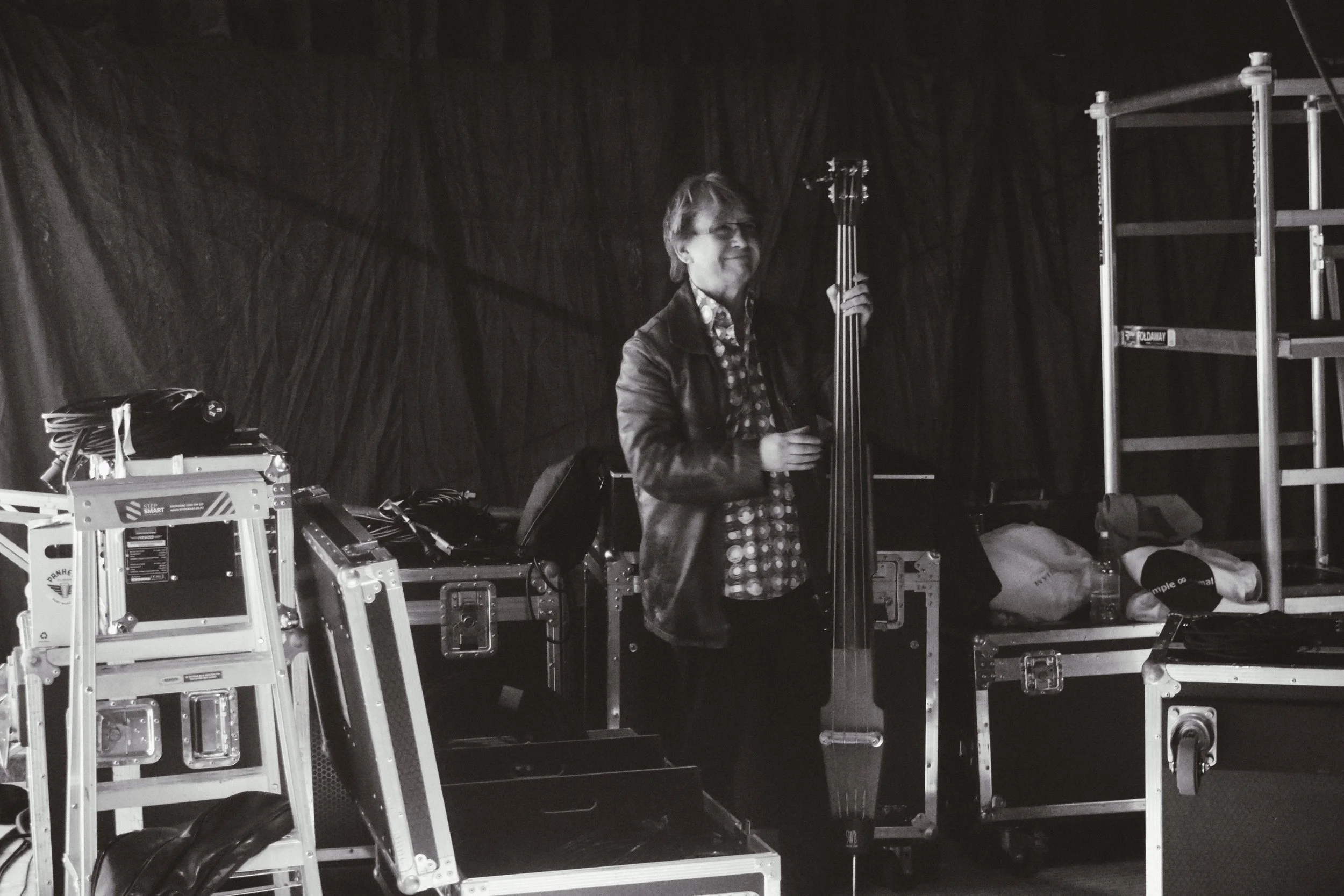 A man holding a double bass on a stage with equipment and storage cases around him.