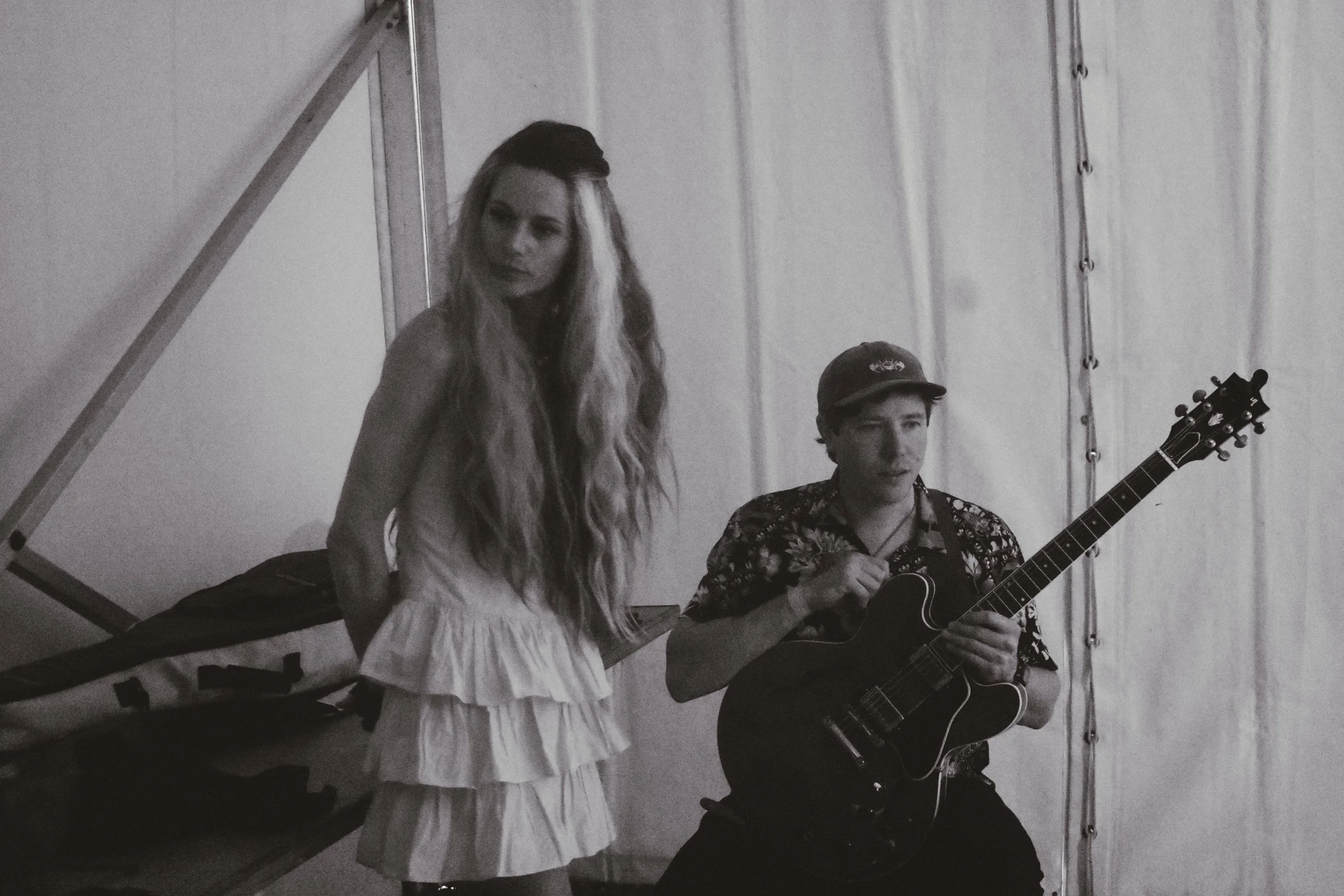 A woman with long, wavy hair wearing a sleeveless dress with ruffles, standing beside a man playing an acoustic guitar, in front of a plain background.