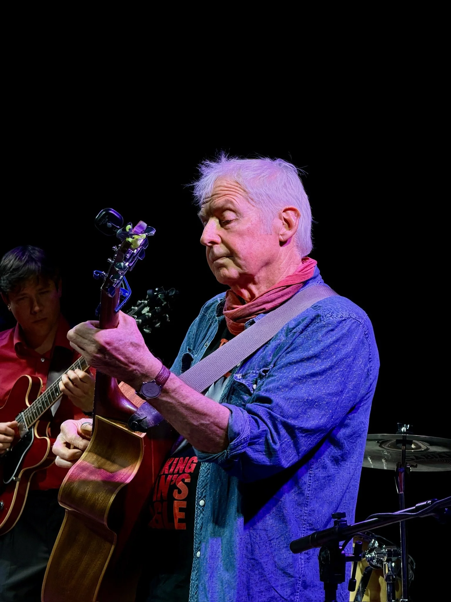 An elderly man with white hair playing an acoustic guitar on stage, accompanied by a younger man with dark hair playing a guitar, against a dark background.