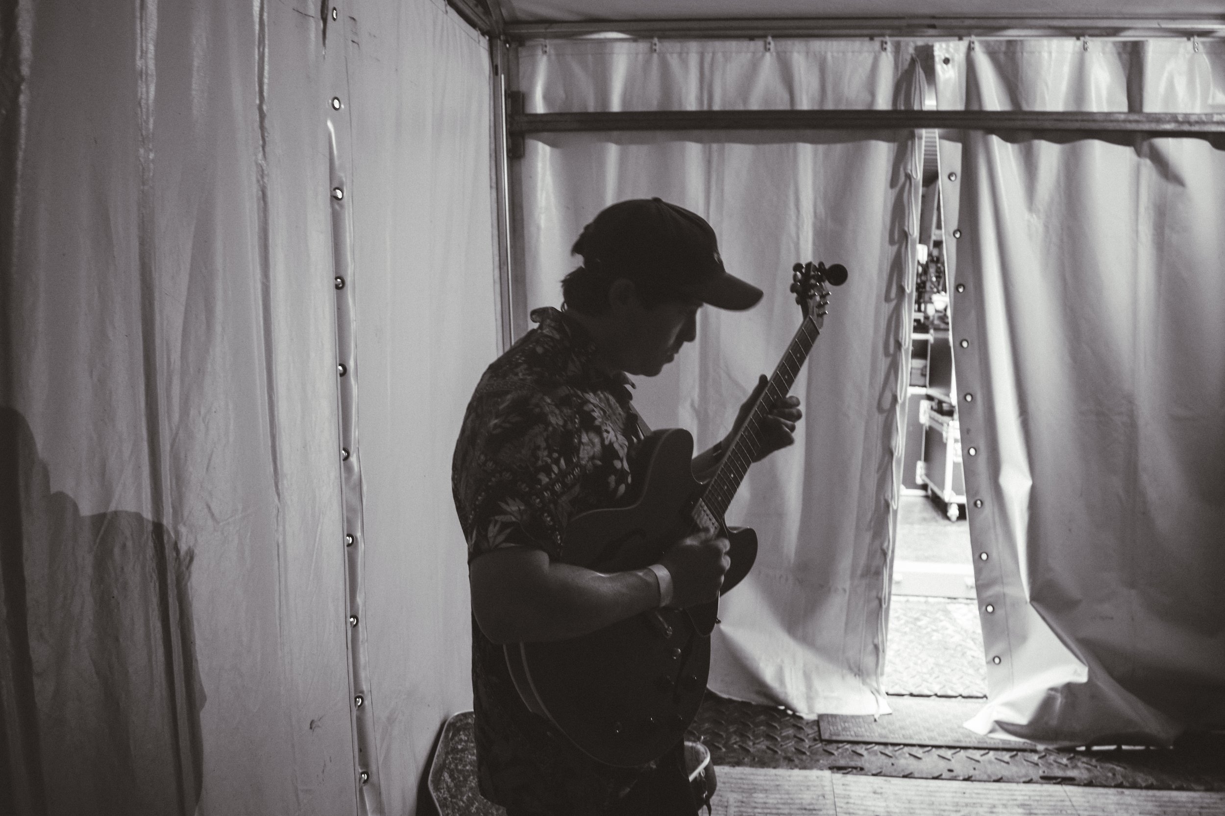 A silhouette of a person wearing a cap and a floral shirt, playing a guitar in a backstage or tent area.