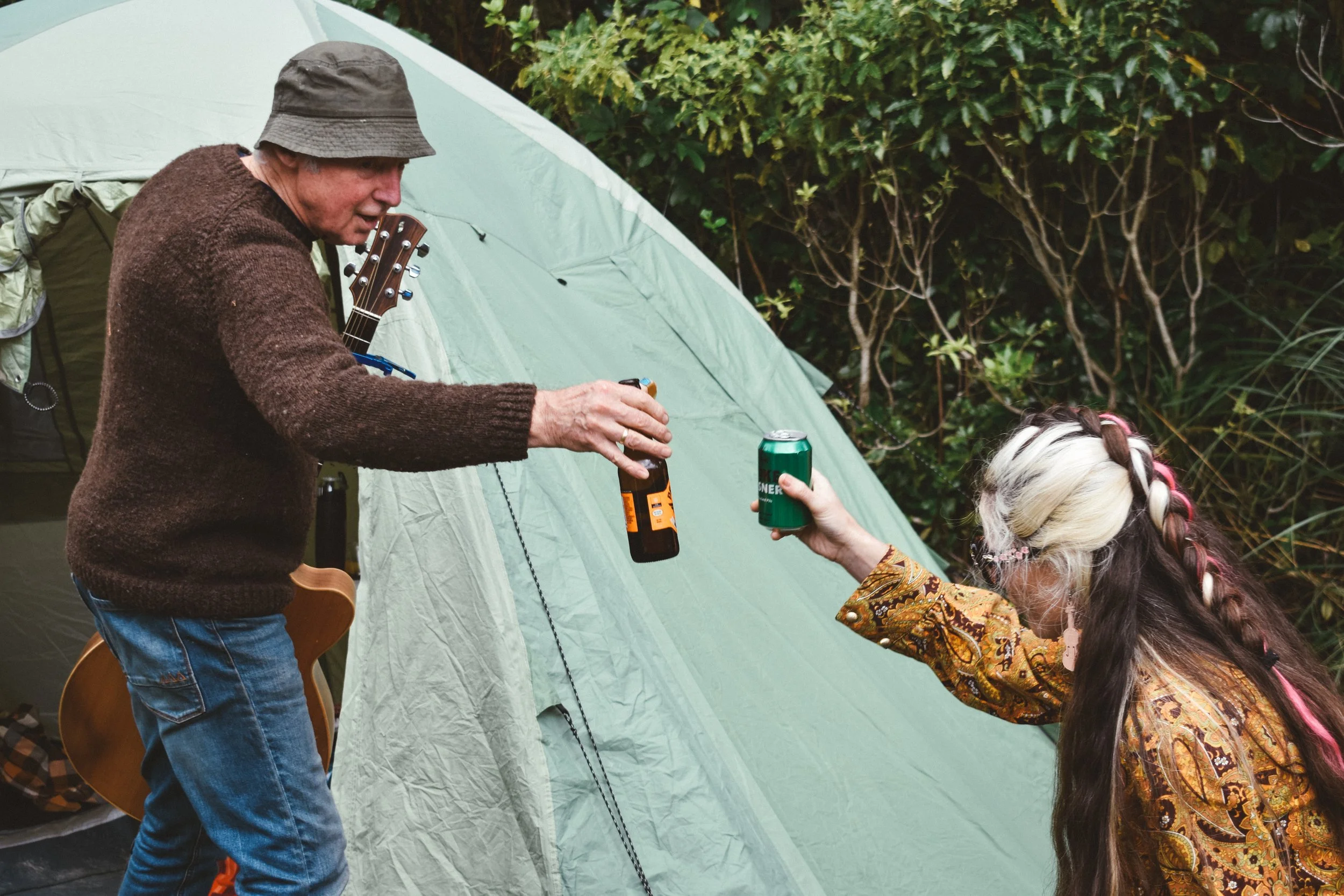 An elderly man wearing a brown sweater and hat standing next to a green camping tent, handing a drink to a young woman with long, braided hair and glasses, in an outdoor setting with greenery.