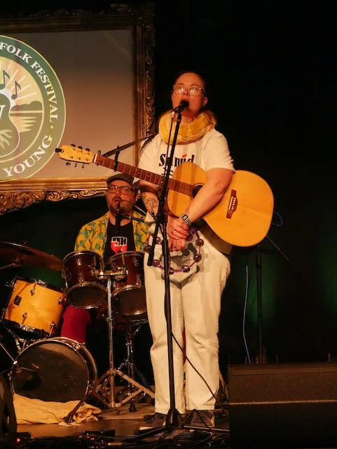A woman with glasses, wearing a yellow scarf and white outfit, holding an acoustic guitar and singing into a microphone on stage. A man with glasses, wearing a colorful shirt, is playing drums in the background. There is a logo on the wall that reads