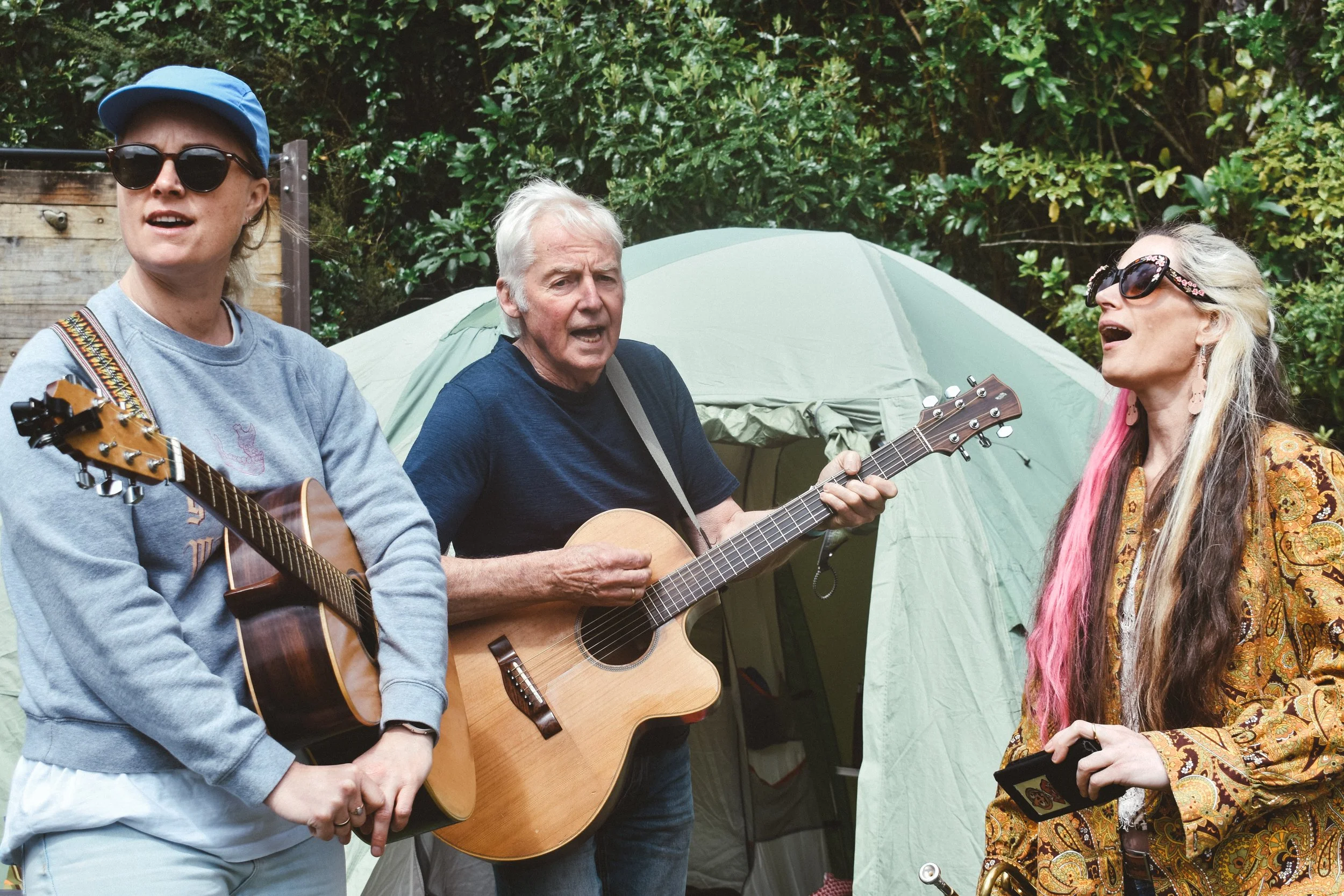 Three people singing and playing guitars outside near a tent, with a background of green bushes and trees.
