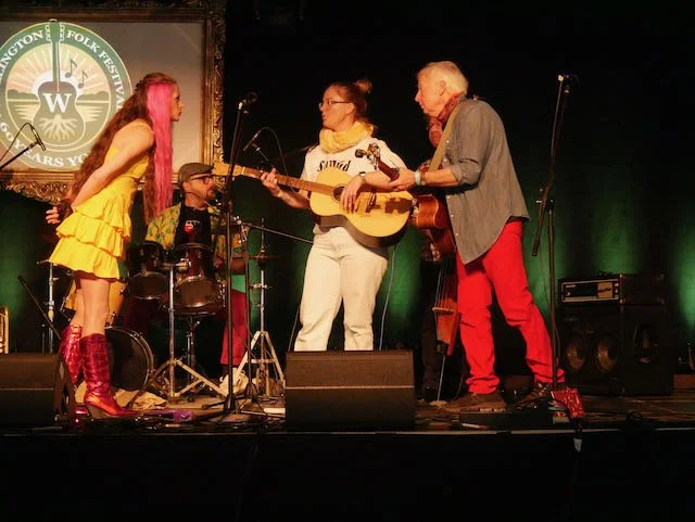 Four musicians performing on stage, with two women and two men, playing guitars and drums at the Wilton Folk Festival.
