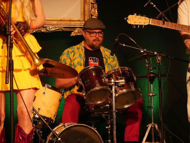 A man playing the drums during a musical performance, with parts of other musicians visible. The man is wearing glasses, a flat cap, a colorful shirt, and red pants.