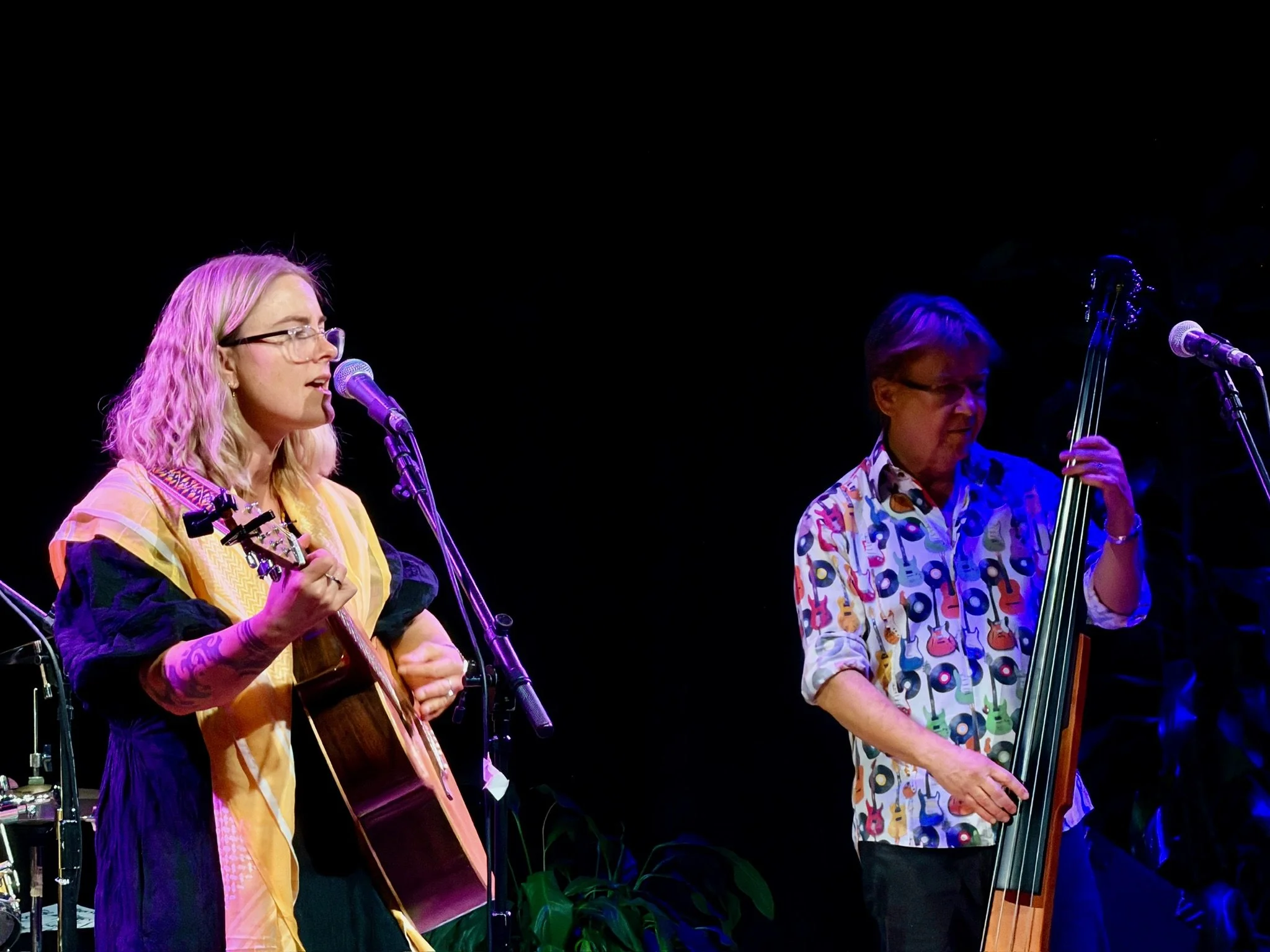 Musicians on stage performing; woman singing and playing guitar, man playing upright bass, both wearing colorful clothing, illuminated by stage lights.