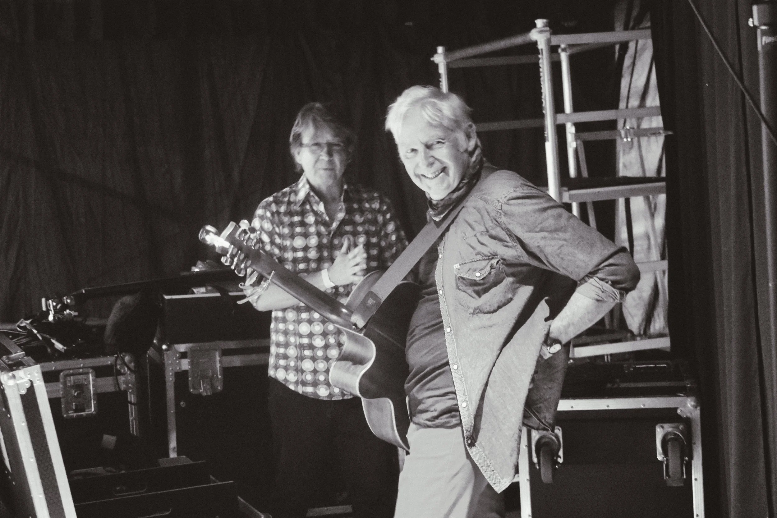 Two men in a music studio, one holding a guitar and smiling at the camera, the other standing behind with a neutral expression, surrounded by musical equipment and stage scaffolding.